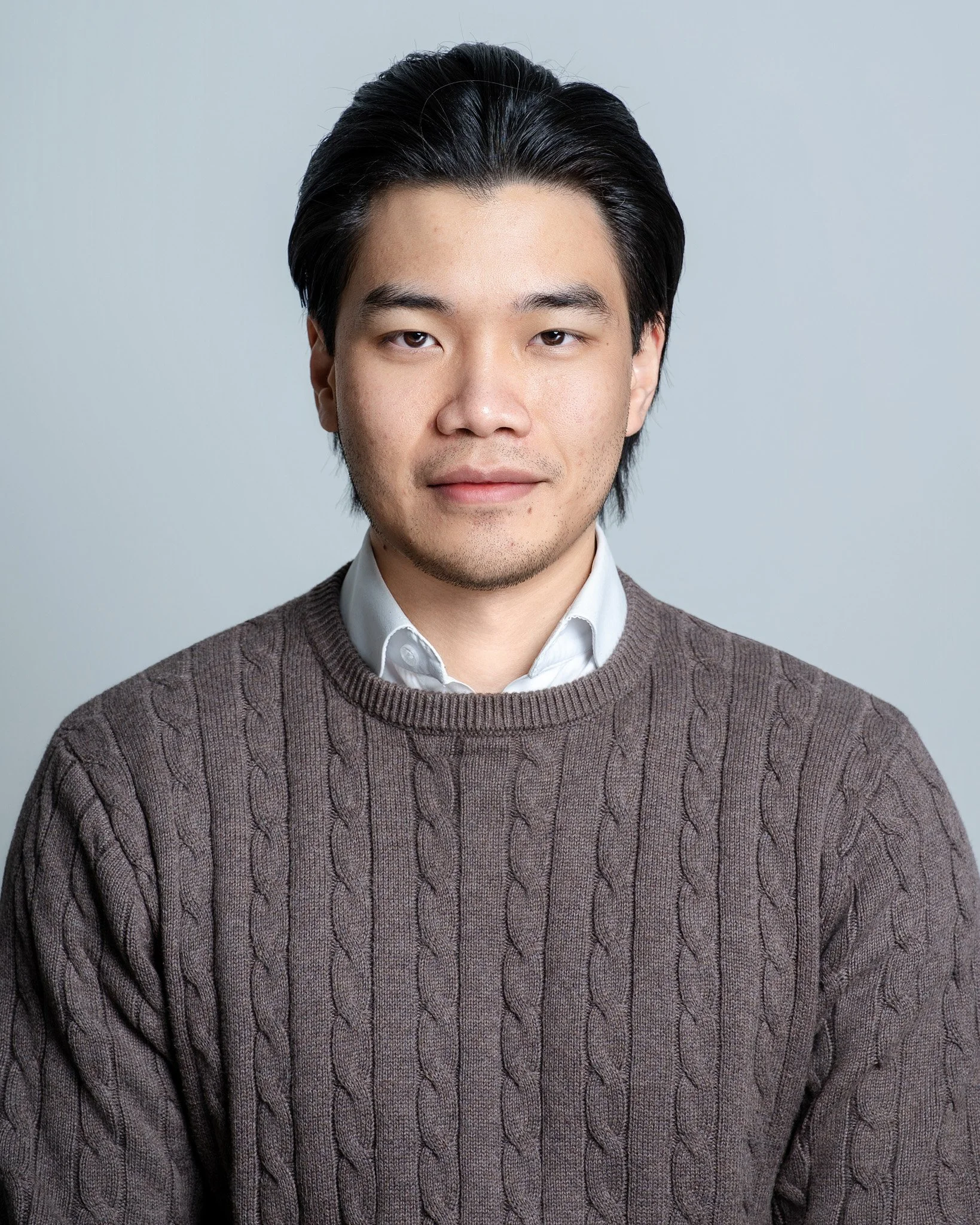 Portrait of a young Asian man with black hair, wearing a white collared shirt under a brown cable-knit sweater, posing against a light gray background.