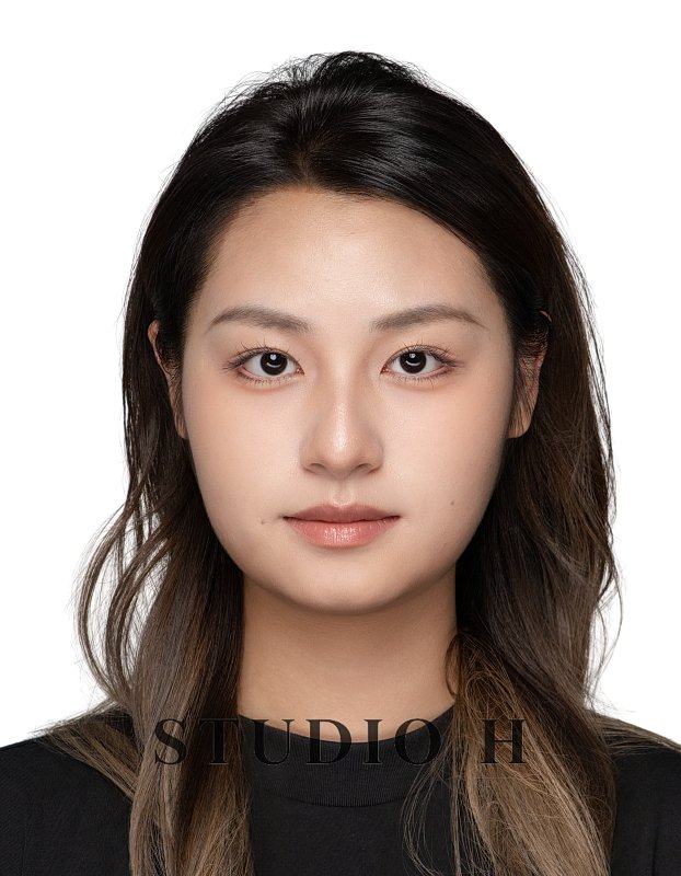 Portrait of a young woman with long wavy dark hair, wearing a black top, looking directly at the camera with a neutral expression, against a plain white background.