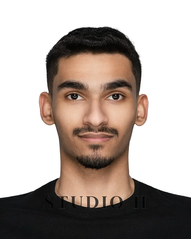 A young man with short dark hair, light facial hair, and a slight smile, wearing a black shirt, facing forward against a white background.