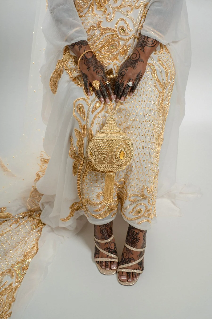A woman wearing a white and gold embroidered dress with gold accessories, holding a gold embroidered clutch, with henna-decorated hands and gold strappy high heels.