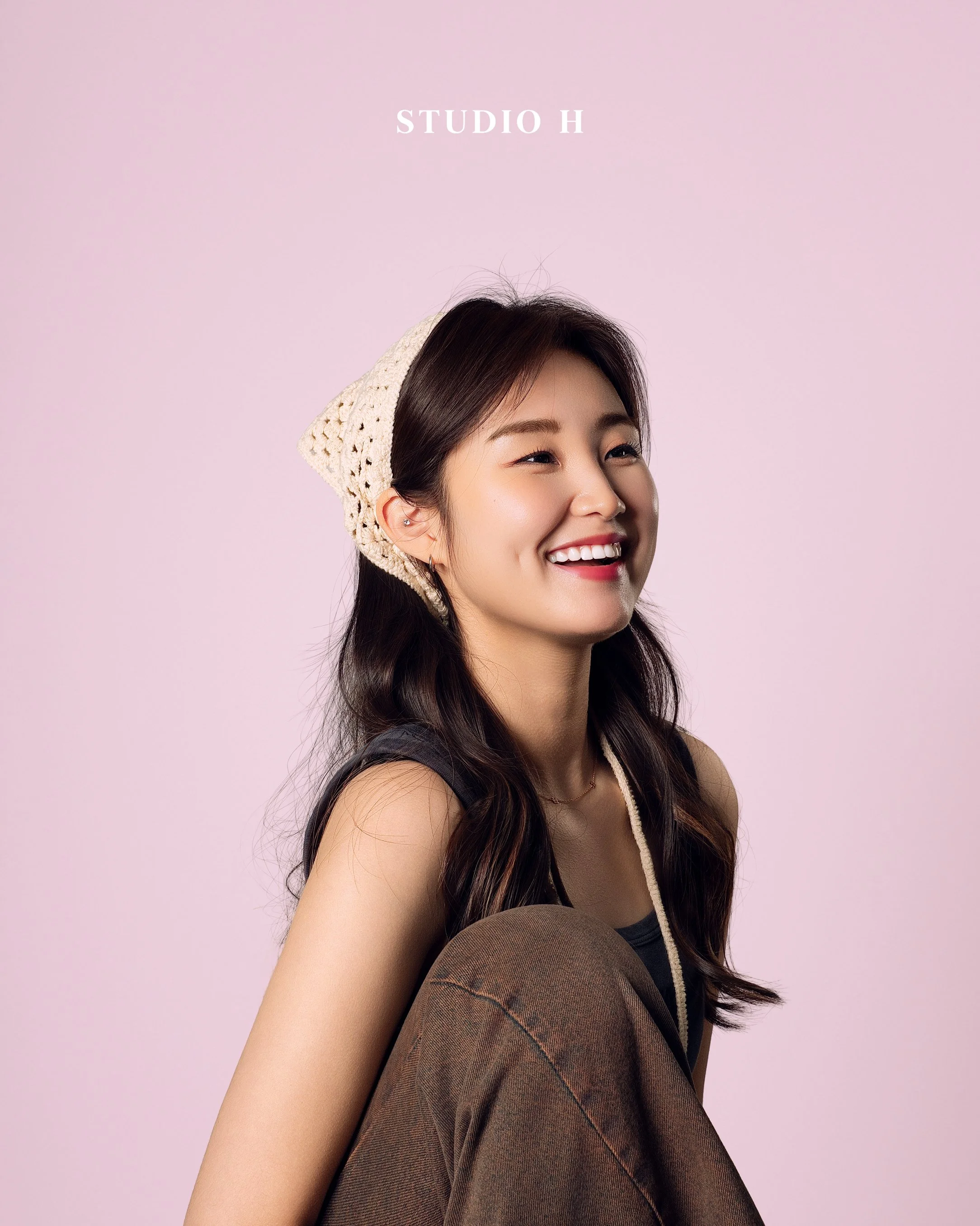 A young woman smiling with long dark hair, wearing a cream-colored knit hat and a sleeveless dark top, against a pink background with 'STUDIO H' text.