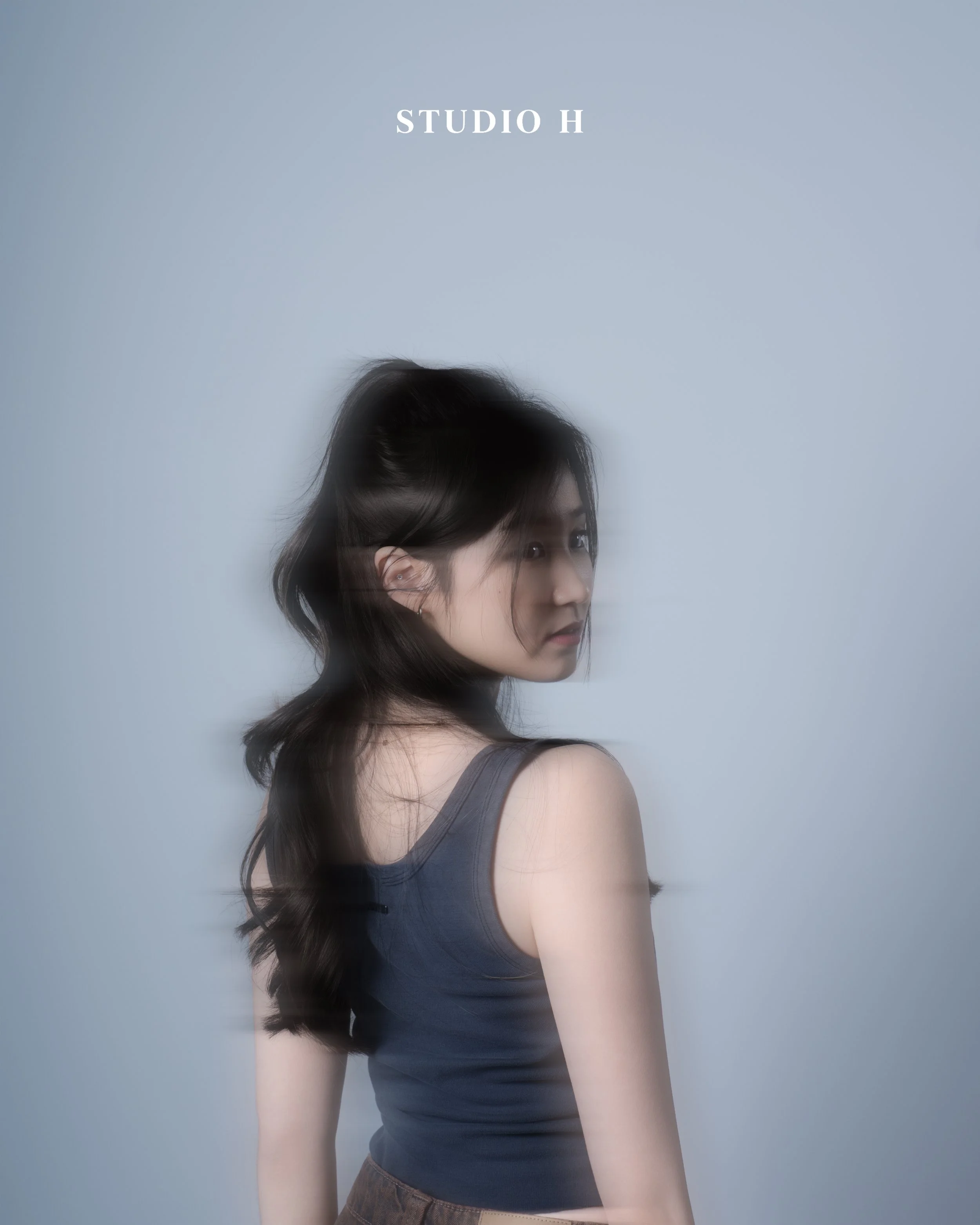 A young woman with long dark hair wearing a sleeveless gray top, looking over her shoulder against a plain light gray background with text "STUDIO H" at the top.