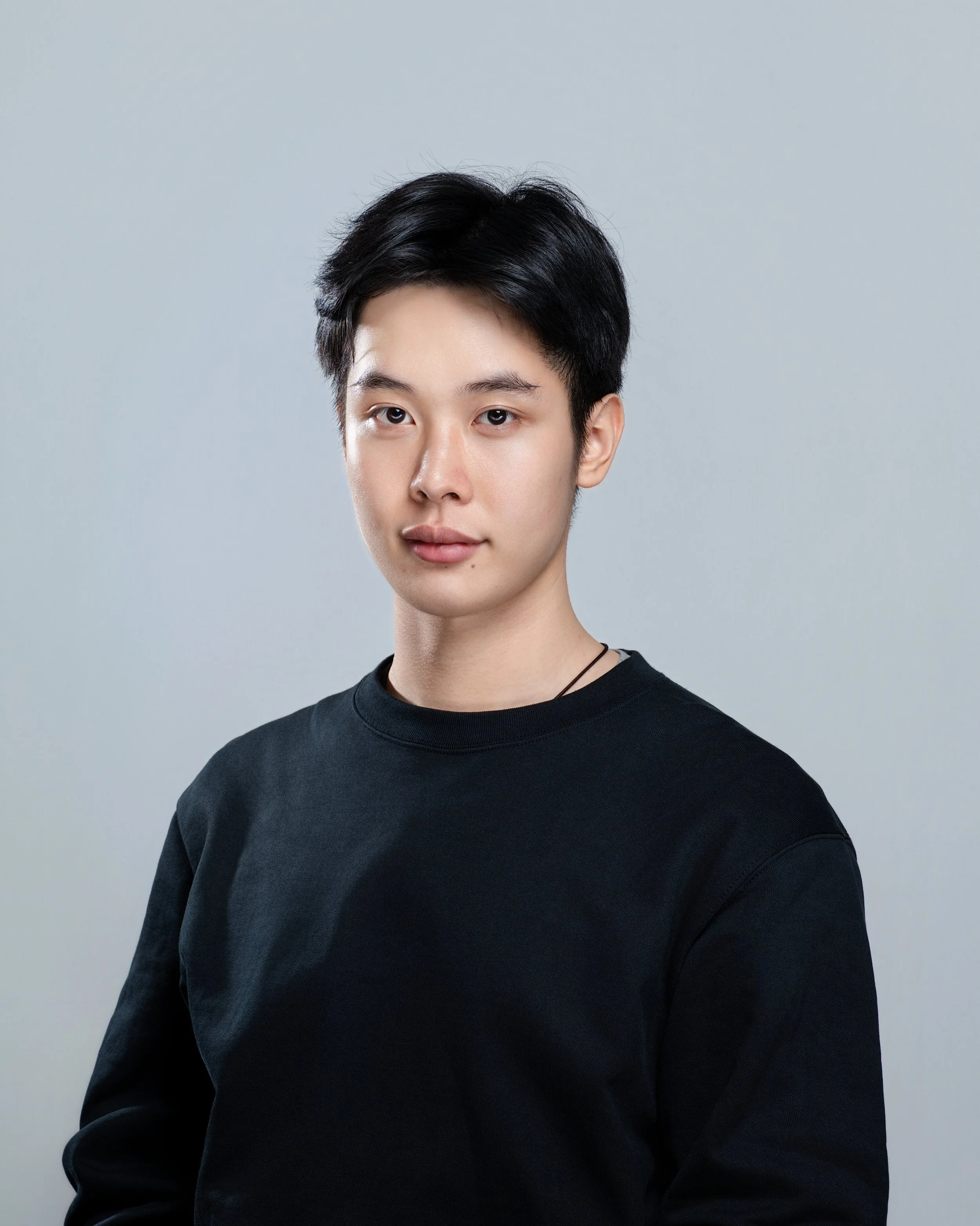 A young person with short black hair, wearing a black shirt, posing against a plain light grey background.