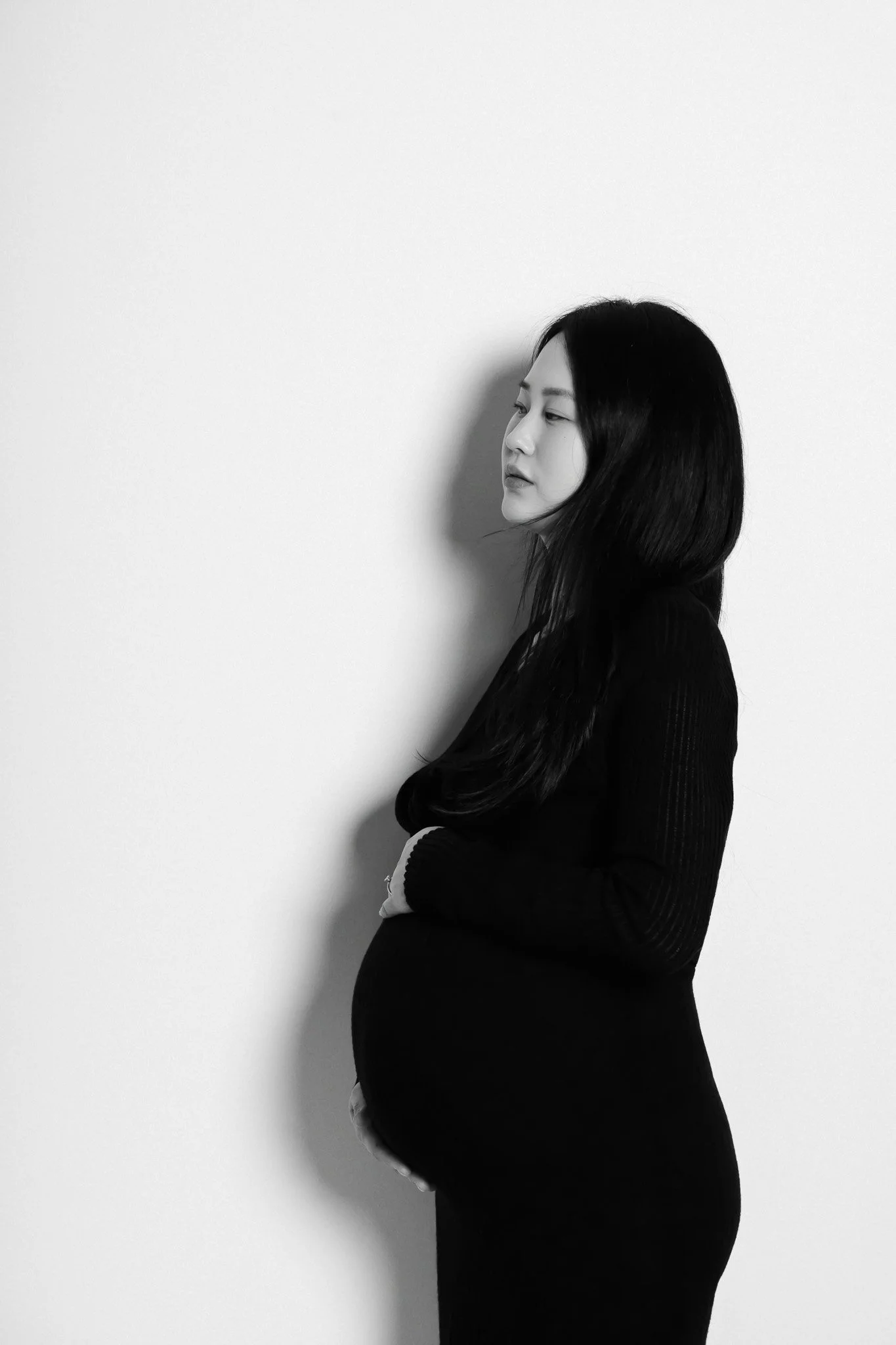 A black and white photo of a woman standing sideways with her arms crossed, wearing a black long-sleeve top, against a plain white wall.