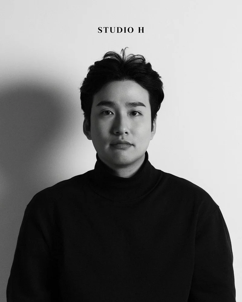 Black and white portrait of a young man with dark hair, wearing a black turtleneck, standing in front of a white wall with the text 'STUDIO H' above his head.