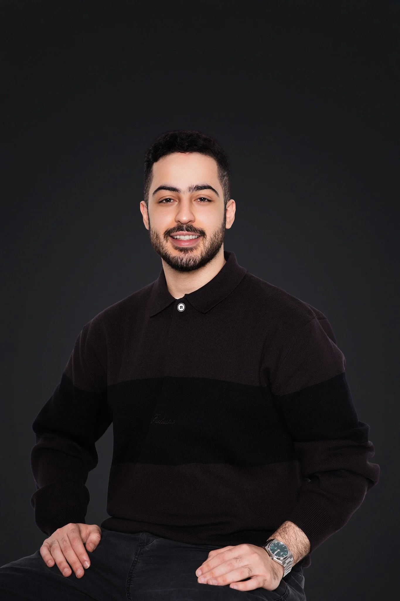 Portrait of a young man with dark hair, beard, and mustache, wearing a black long-sleeve shirt with a collar, sitting against a dark background, smiling and looking at the camera.