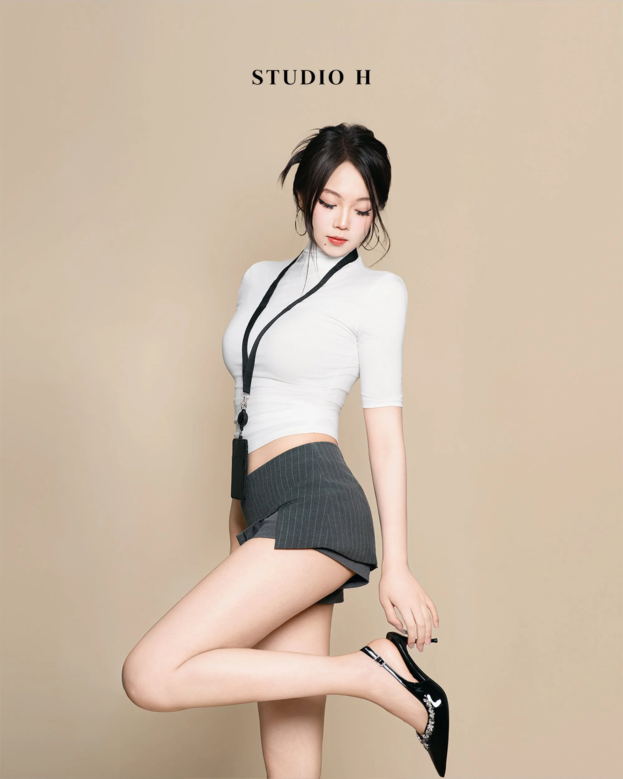 A young woman with black hair in a bun, wearing a white crop top, black pinstripe skirt, and shiny black high heels, standing against a beige background with "STUDIO H" printed above her.