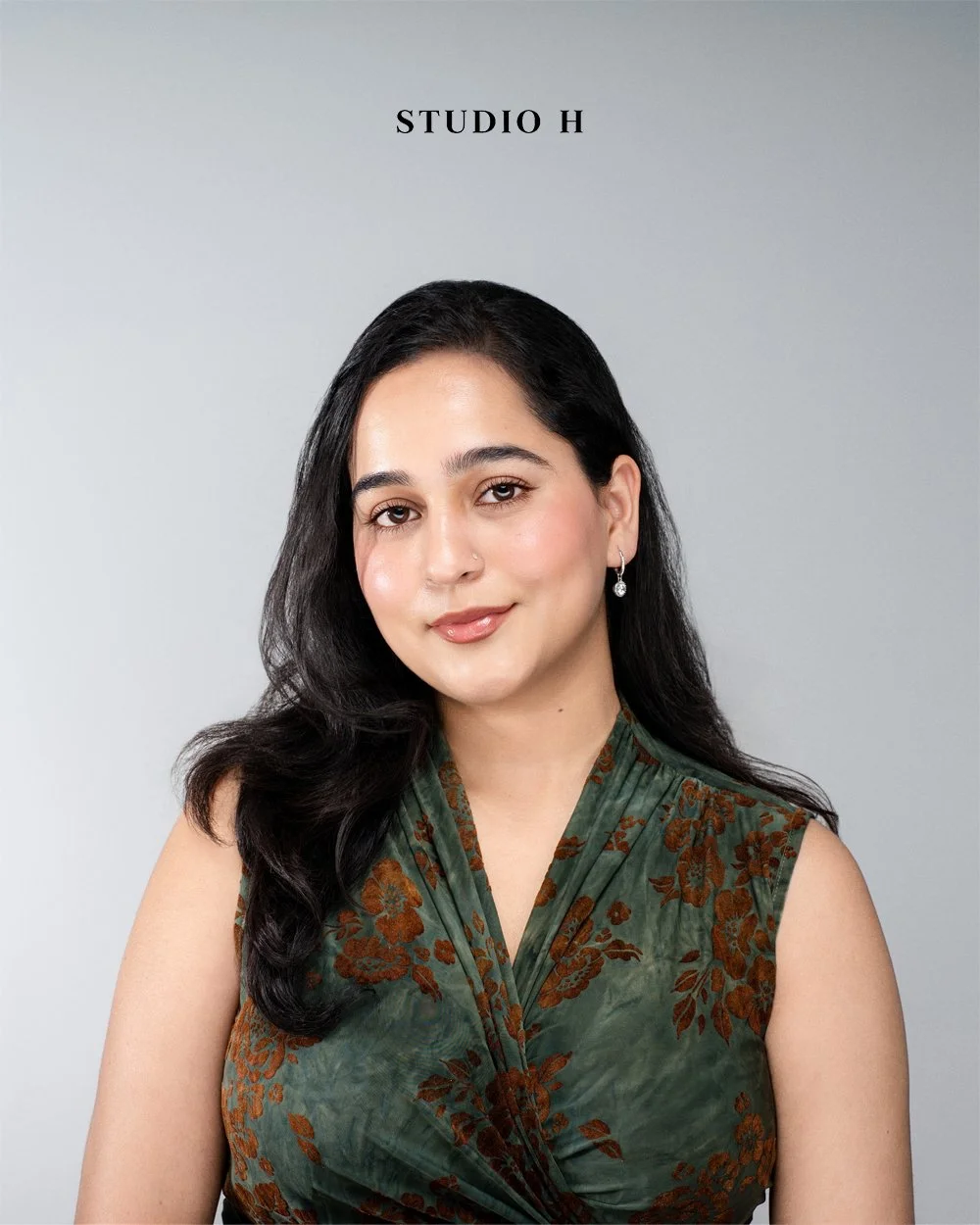 A woman with long black hair, wearing a sleeveless green dress with brown floral patterns, posing in front of a gray background with the words 'STUDIO H' at the top.