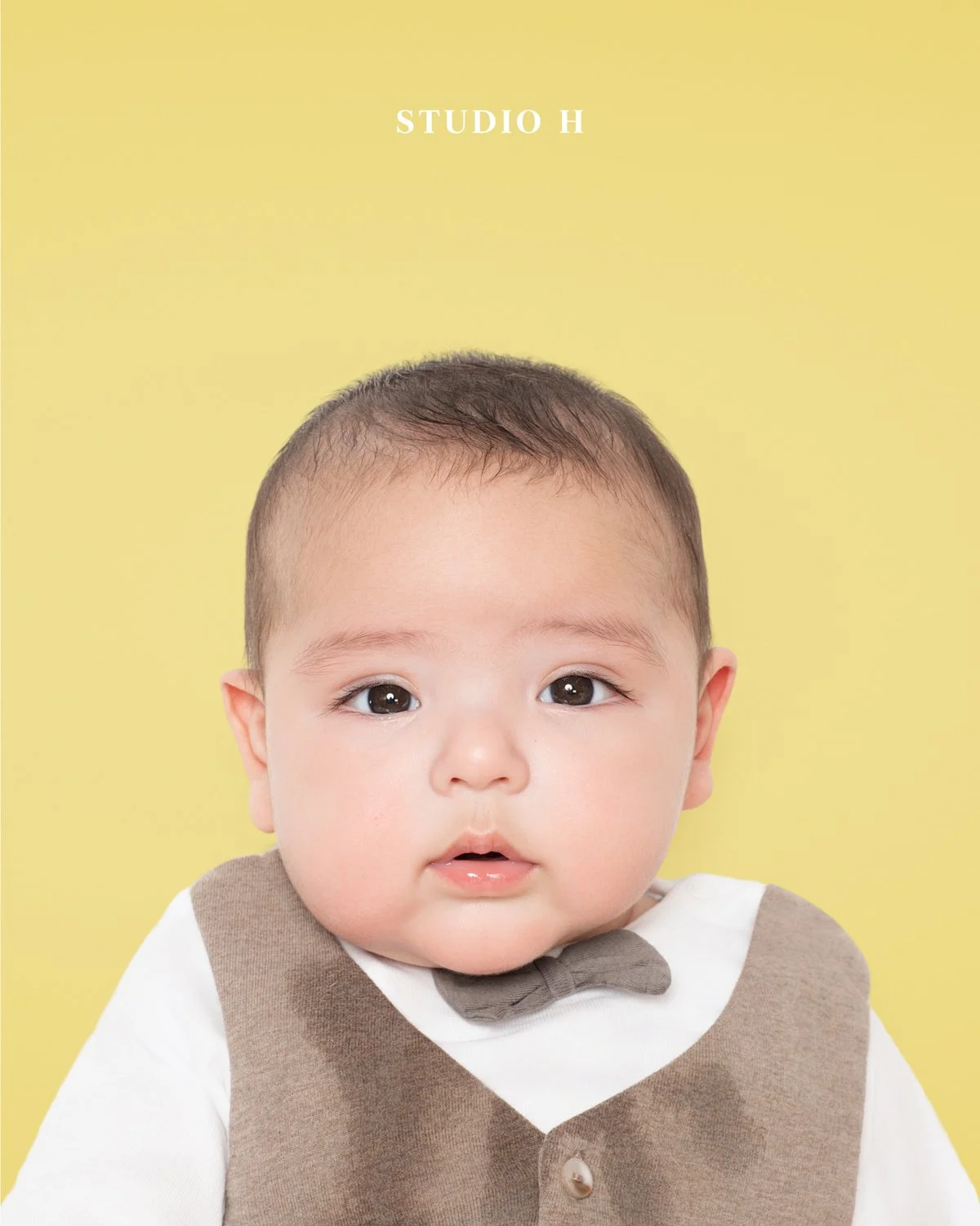 A baby with dark hair and brown eyes wearing a beige vest and white shirt with a brown bow tie, against a yellow background.
