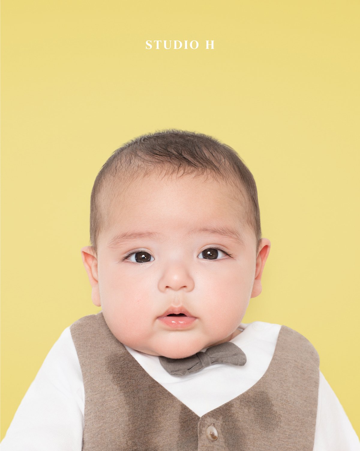 A baby with short dark hair, wearing a white shirt and a brown vest with a bow tie, against a yellow background.