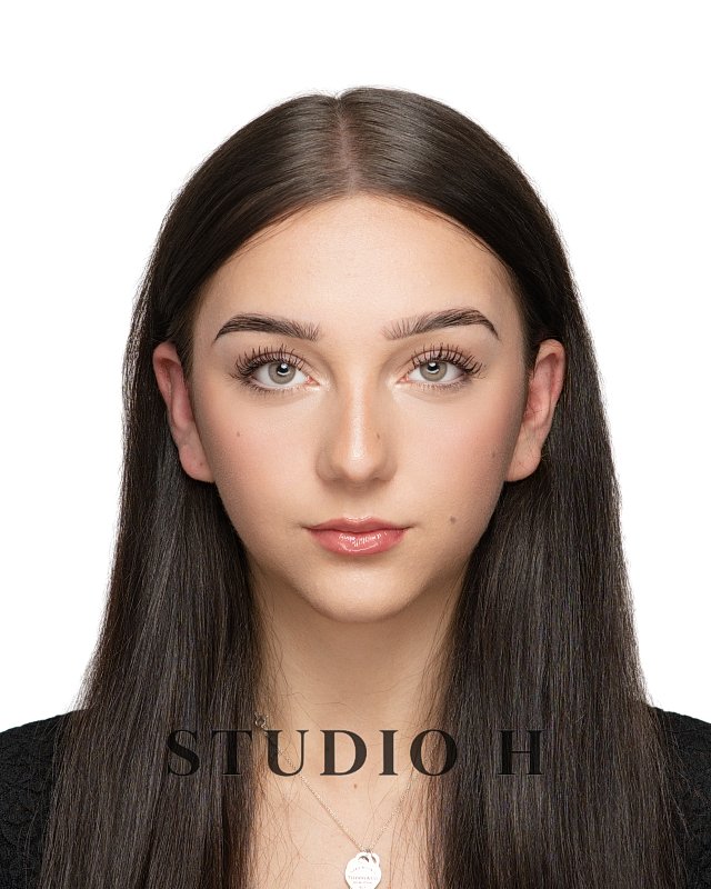 A young woman with long, straight brown hair, wearing a black top and a pendant necklace, looking directly at the camera with a neutral expression, against a white background.