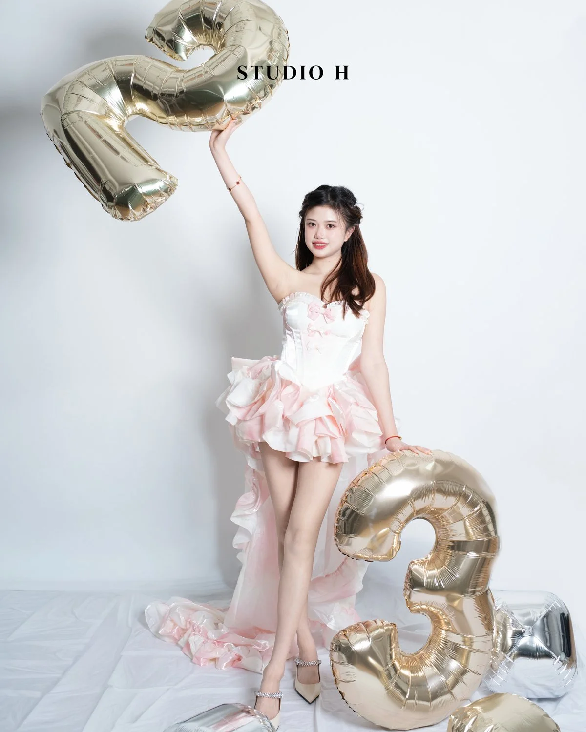 A young woman in a pink and white dress standing in front of a plain white background, holding a large gold balloon shaped as the number 2 in her right hand and a similar balloon in her left hand, with more balloons scattered around her feet.