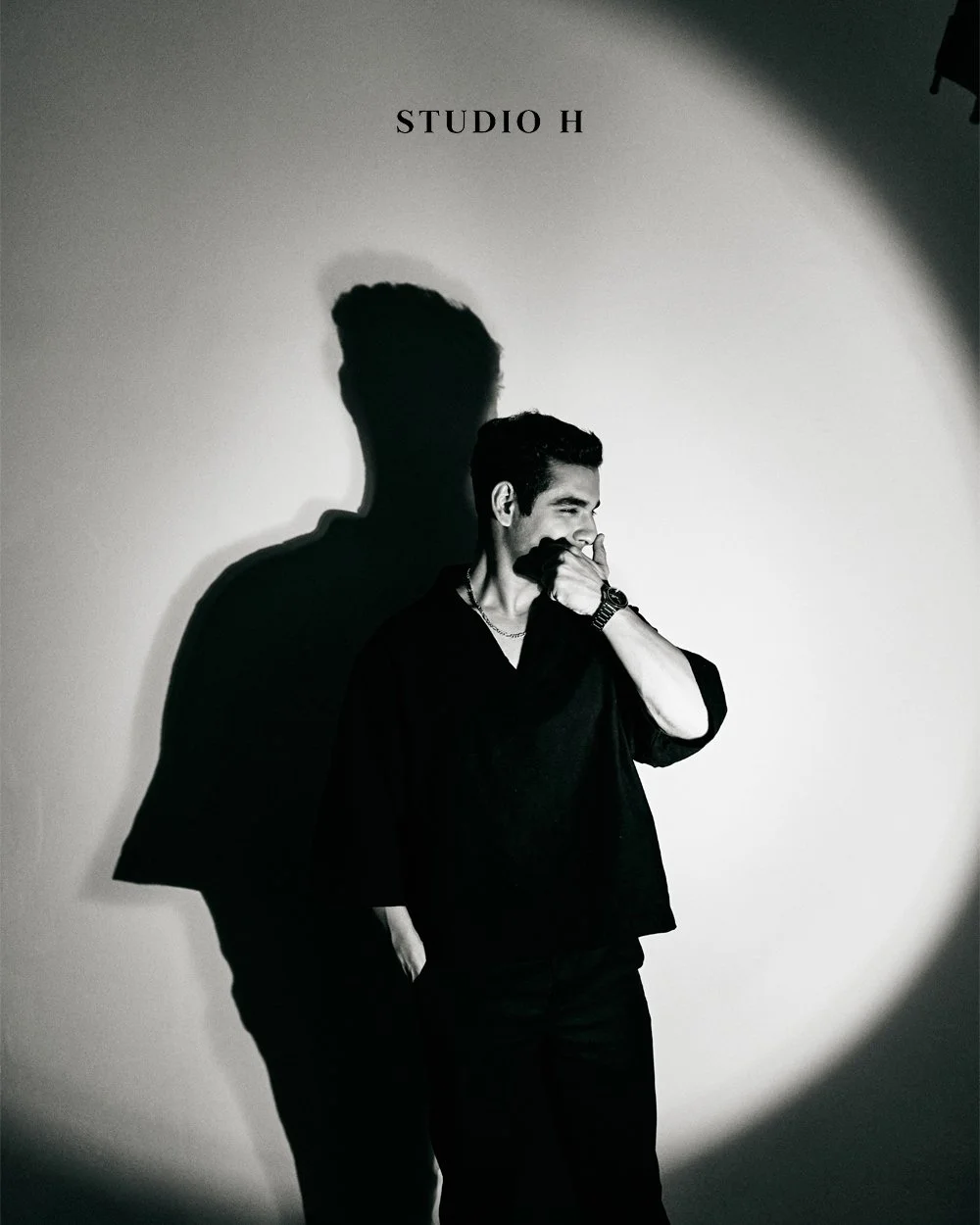 A young man in black clothing standing against a plain wall with a spotlight creating a large shadow behind him. The wall has the words 'STUDIO H' written at the top.