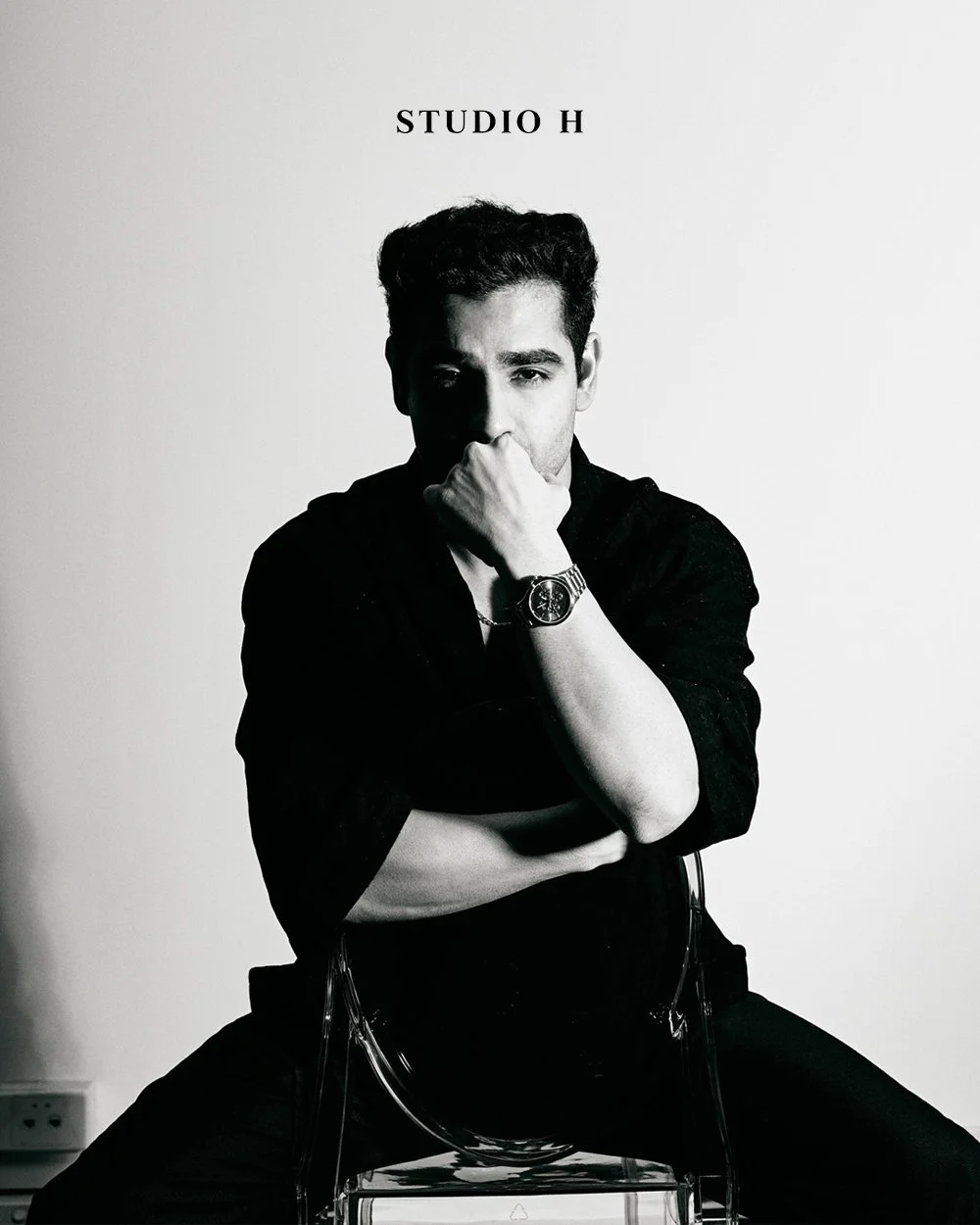Black and white photo of a young man sitting on a transparent chair, dressed in dark clothing and a watch, with his right hand covering his mouth and his left arm crossed over his chest. The background is plain, and the text 'STUDIO H' appears overhe