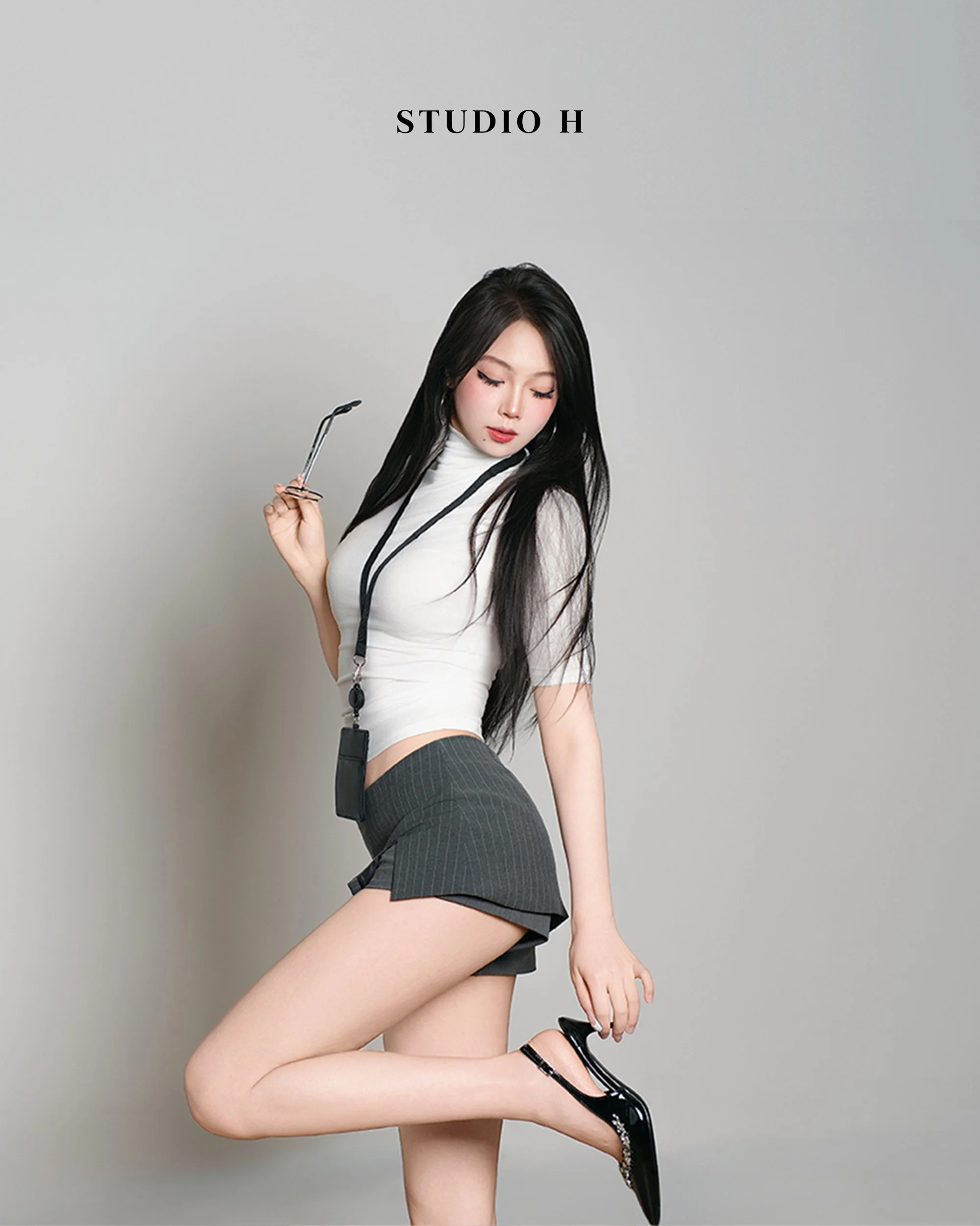 A young woman with long black hair wearing a white top, dark pinstriped shorts, and black high heels, holding glasses in one hand and posing with one leg bent. The background is plain grey with the words 'STUDIO H' above her.