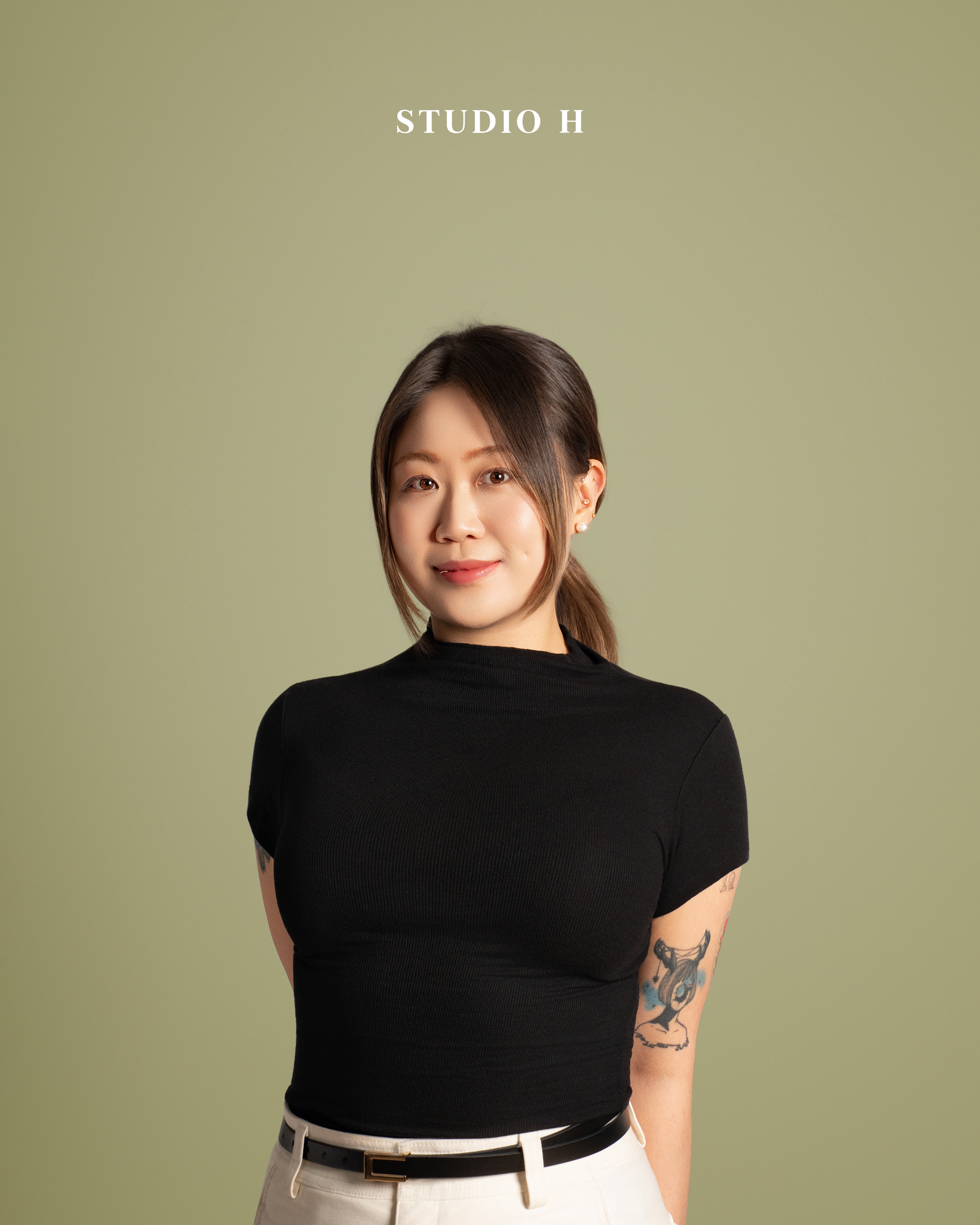 A woman with long dark hair tied in a low ponytail, wearing a black short-sleeve turtleneck shirt and beige pants, standing against a light green background with text that reads "STUDIO H."