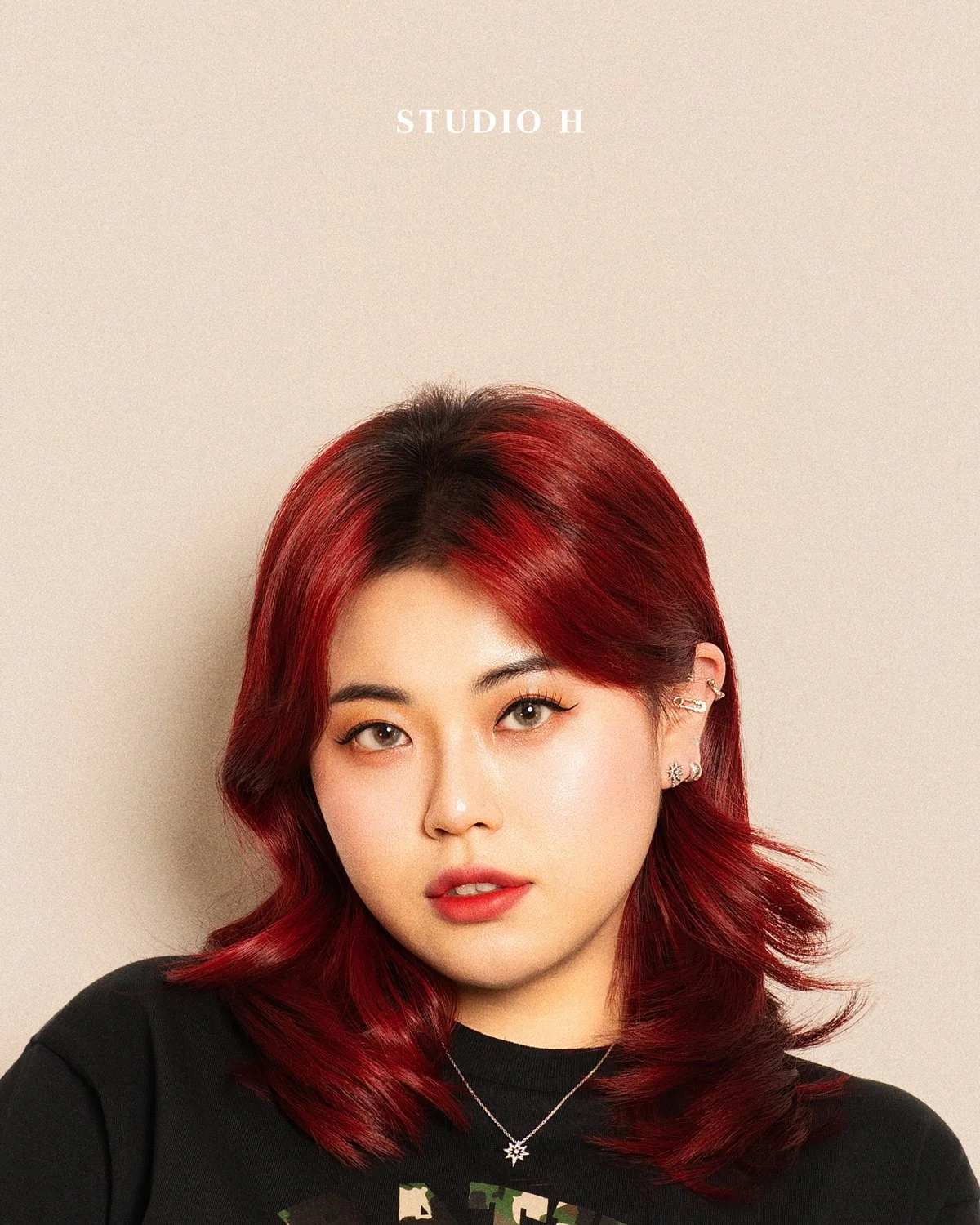 Studio H logo above a woman with red hair styled in loose waves, wearing multiple earrings, a star-shaped necklace, and a black shirt, standing against a plain beige background.