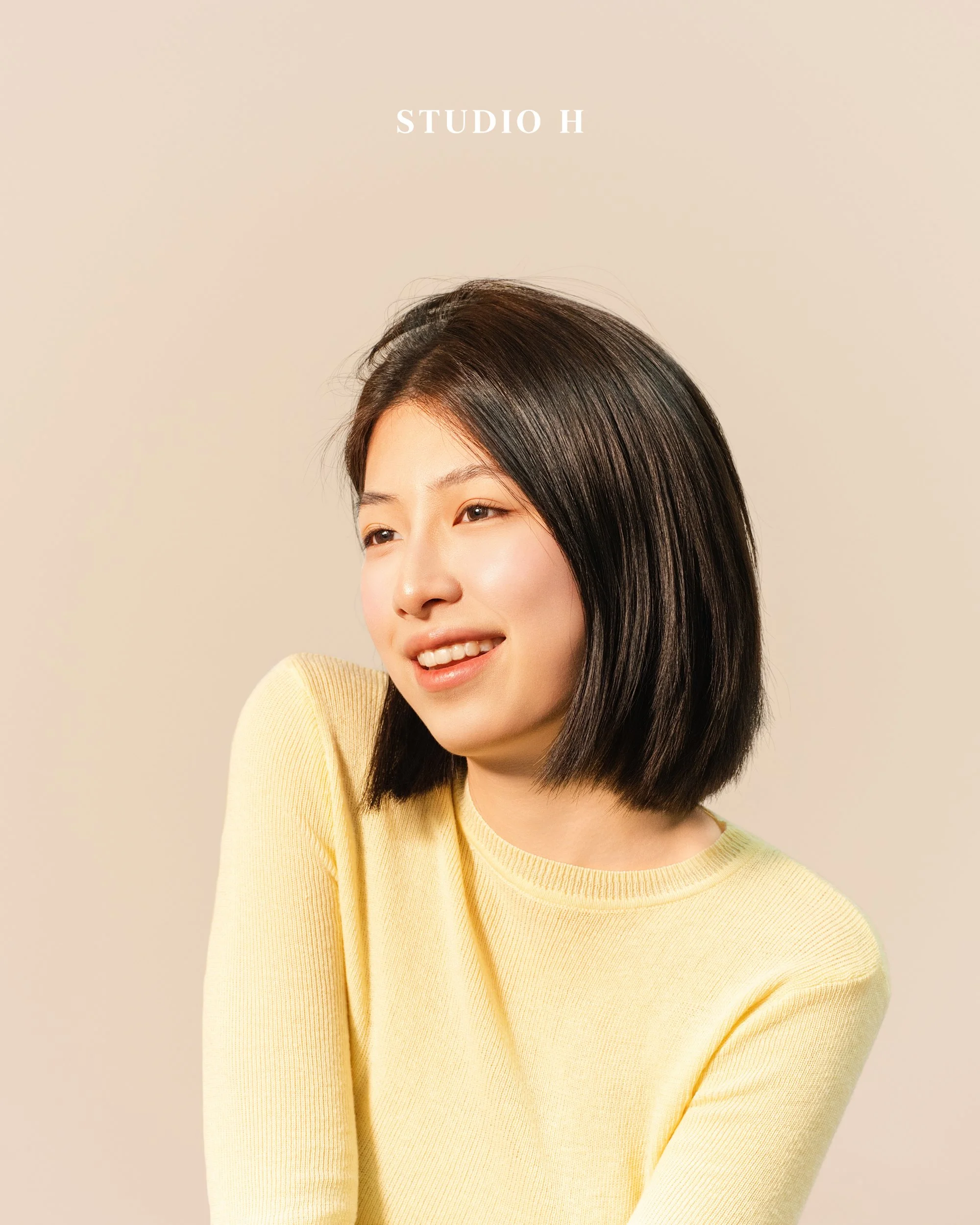 A young woman with shoulder-length black hair smiling and looking to the side, wearing a light yellow long-sleeve shirt, against a plain beige background with ''STUDIO H'' text at the top.