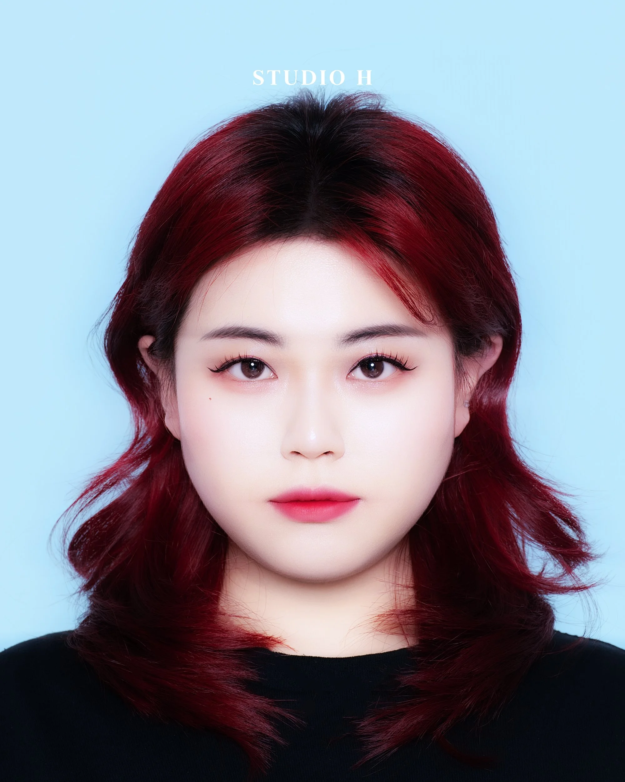 A portrait of a young woman with red hair styled in loose waves, wearing a black top, against a light blue background. Text at the top reads "STUDIO H."