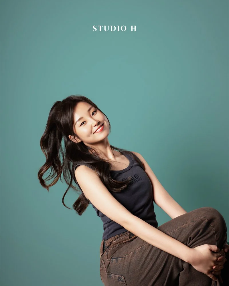 Young woman with long dark hair smiling, sitting with a knee bent, against a plain teal background with the text "STUDIO H" at the top.