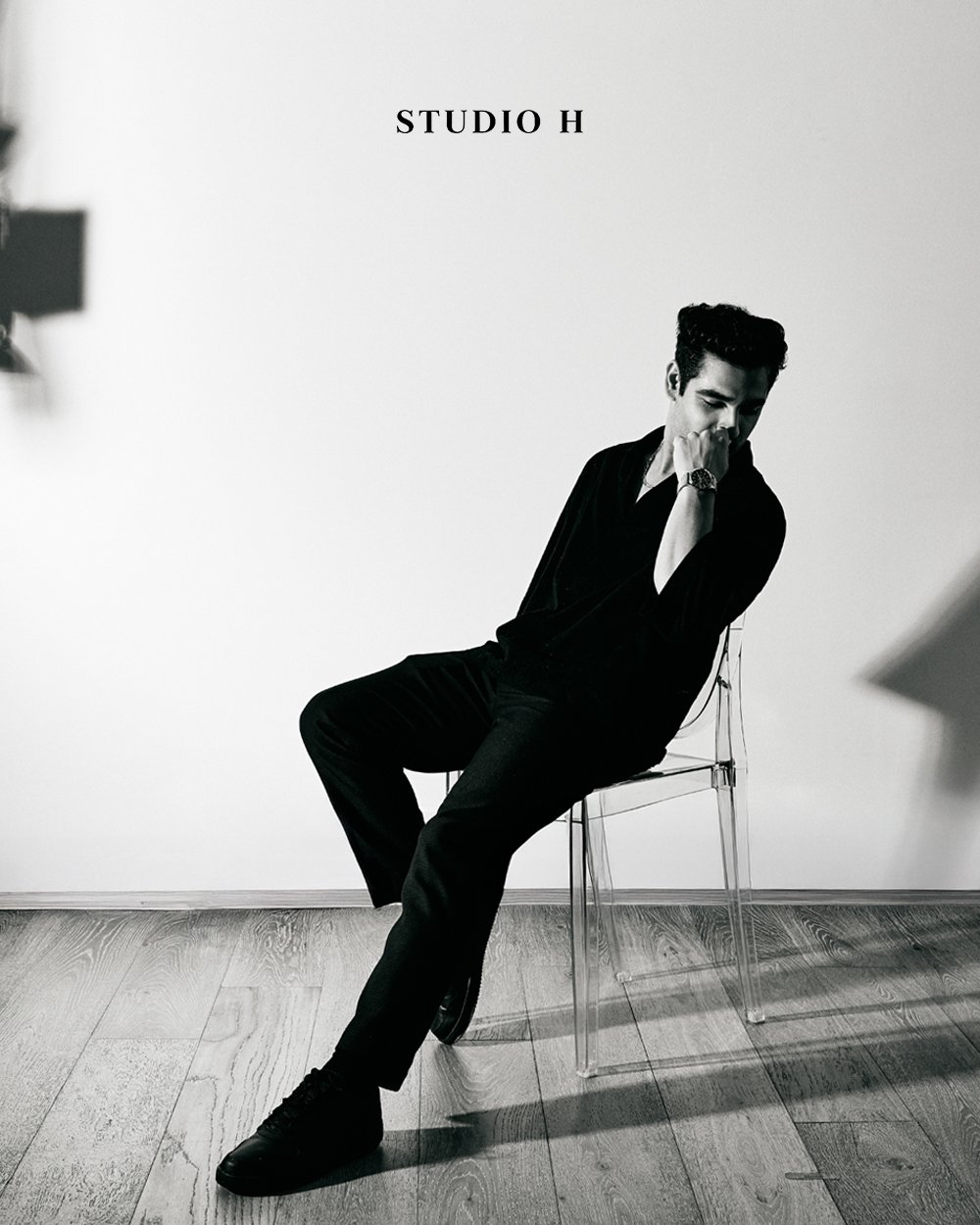 Black and white photo of a man sitting on a transparent chair, looking down with his hand near his face, wearing dark clothing. The background is plain with the words 'STUDIO H' printed on the wall.