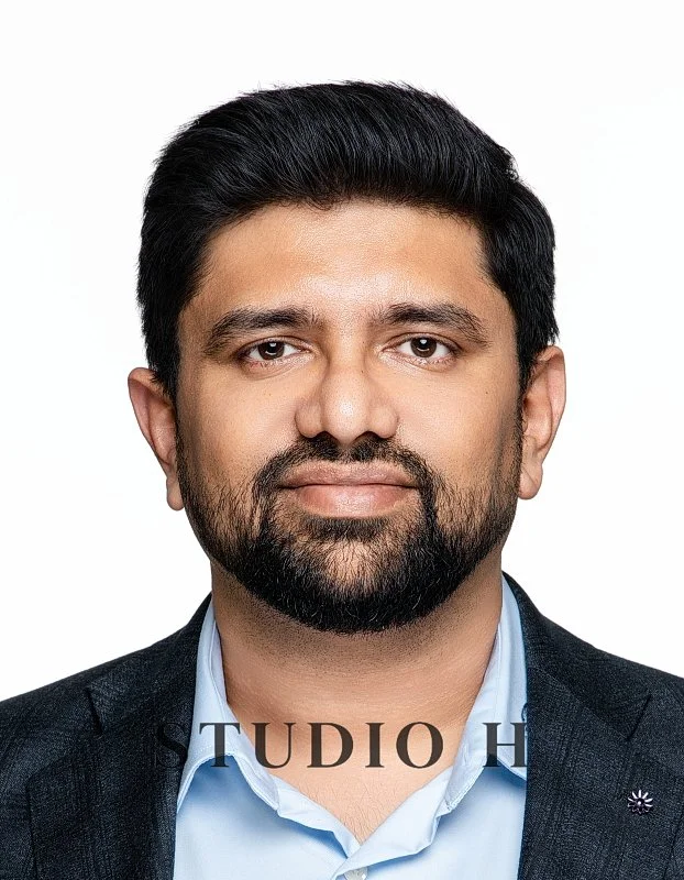 A professional portrait of a man with dark hair and a beard, wearing a suit jacket and light blue shirt, against a plain white background.