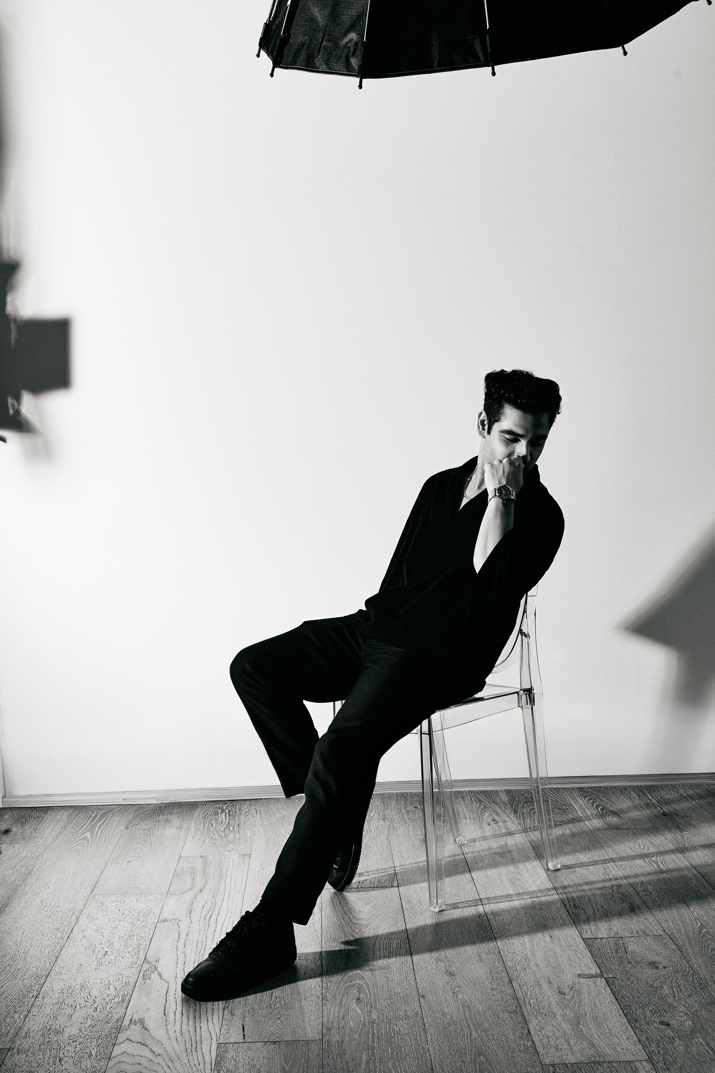 A black-and-white photo of a man sitting on a transparent chair in a studio setting, with a plain white background. He is dressed in dark clothing and has a watch on his left wrist, with his hand resting near his face, looking down thoughtfully.