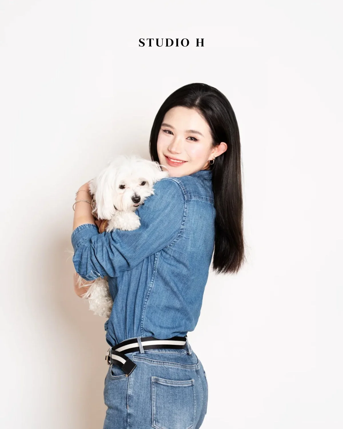 A woman with long dark hair, wearing a denim shirt and jeans, holding a small white dog and smiling, against a plain white background with the text 'STUDIO H' above.
