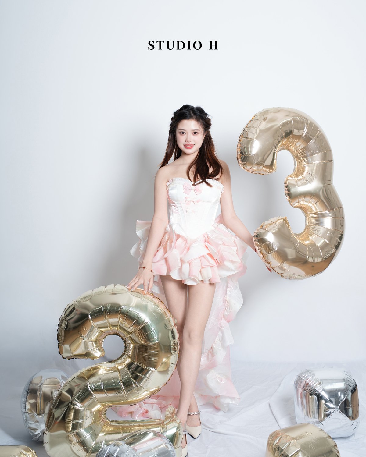 A young woman in a pink dress holding number 3 balloons, standing against a white background with the text 'STUDIO H' above her.