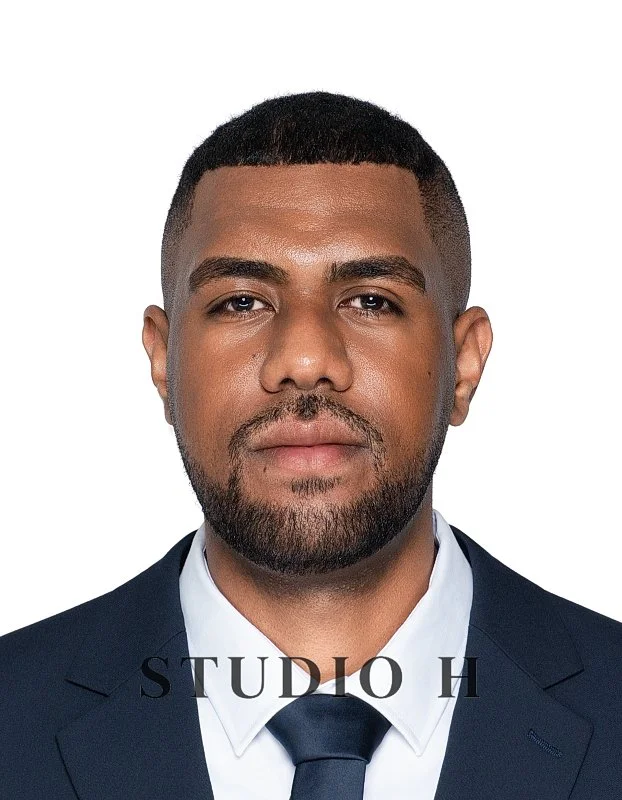 A professional headshot of a man in a dark suit, white shirt, and dark tie, with a serious expression and short hair, against a white background with the text 'STUDIO H'.