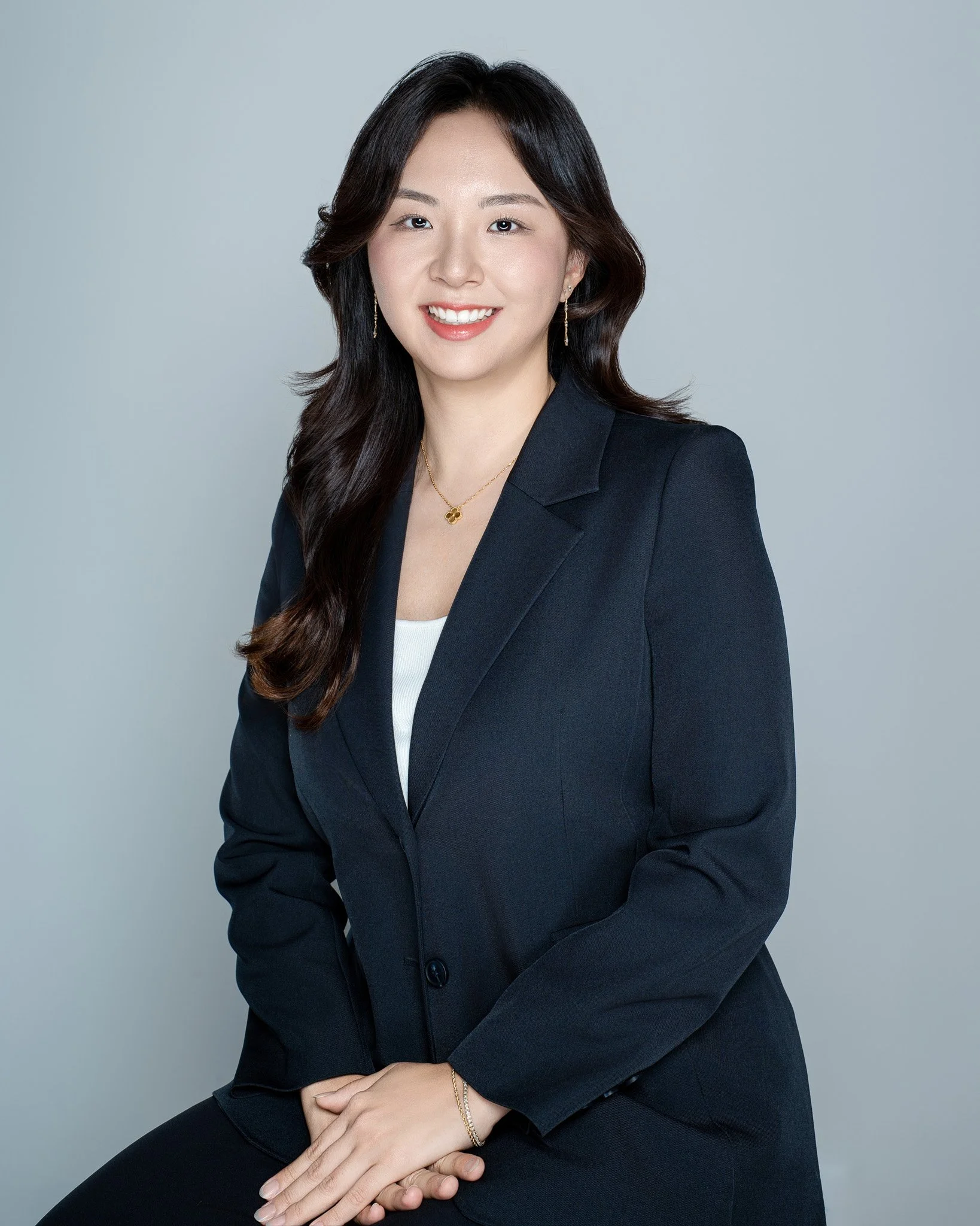 Professional portrait of a woman with long dark hair, wearing a black blazer, white top, gold jewelry, and smiling against a gray background.
