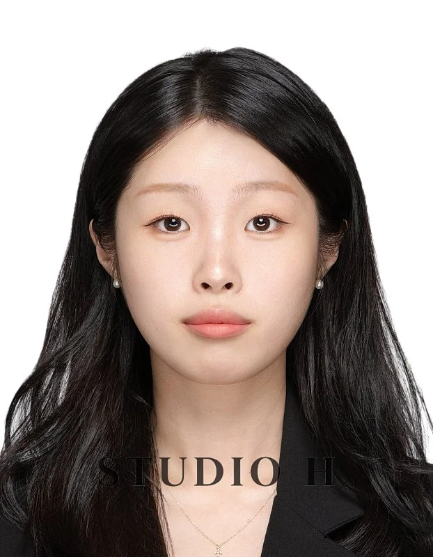 Portrait of a young woman with long black hair, wearing pearl earrings and a black top, against a plain white background.