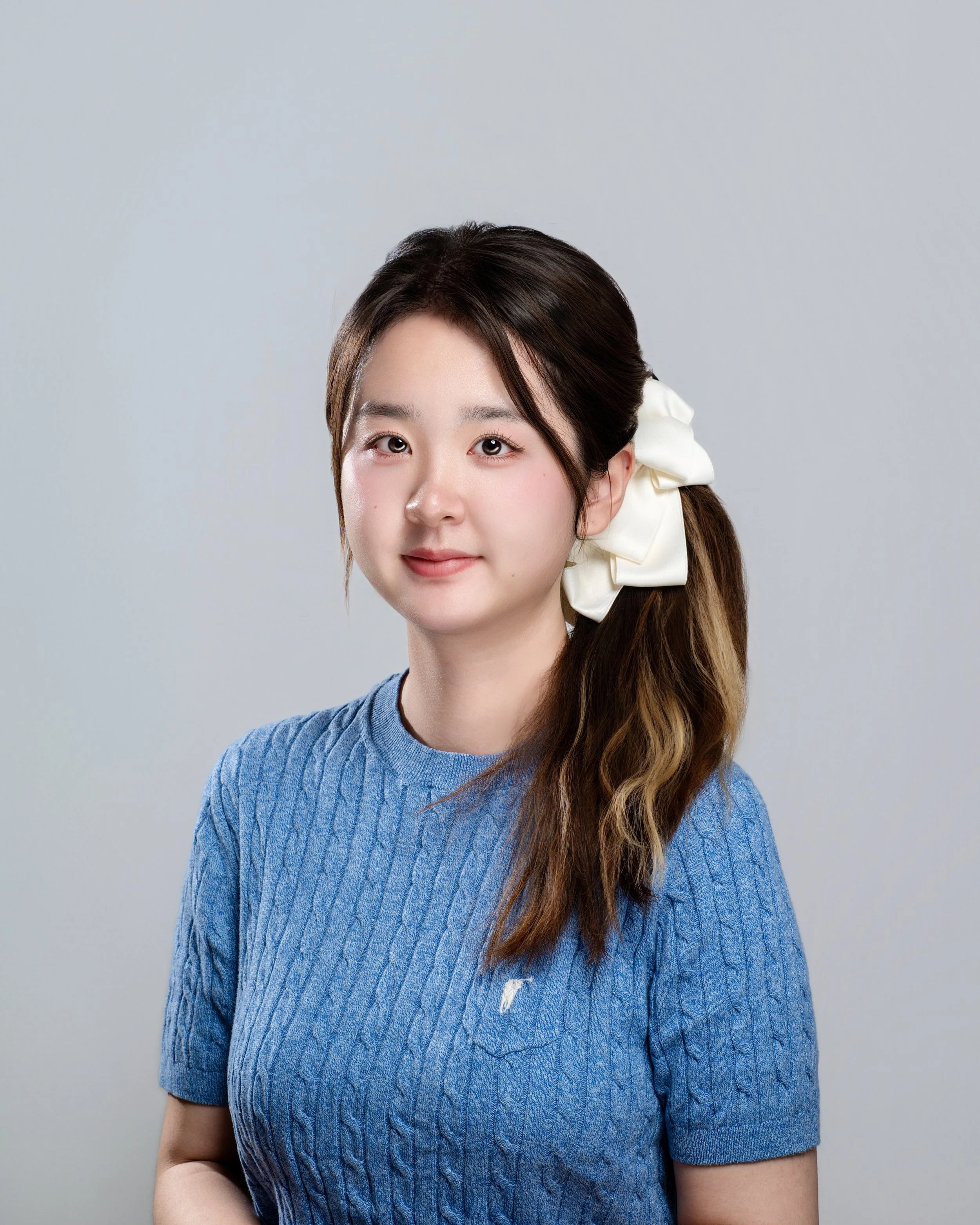 A young woman with dark brown hair, tied to the side with a large white bow, wearing a blue sweater standing against a plain light gray background.