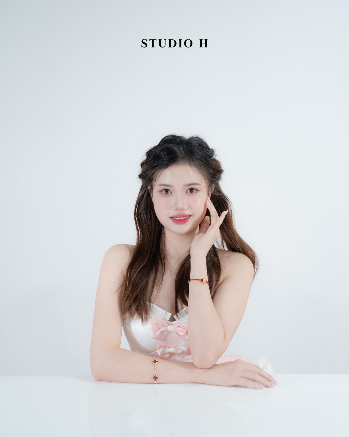 A young woman with long brown hair, wearing a white dress with pink ribbons, sitting with her arms resting on a white surface, touching her face slightly, against a plain white background with 'STUDIO H' text at the top.