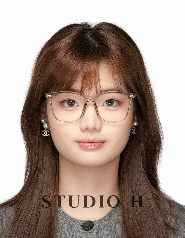 A woman with straight brown hair, wearing clear glasses and Chanel earrings, looking directly at the camera with a neutral expression, against a plain white background. The text 'STUDIO H' is at the bottom of the image.
