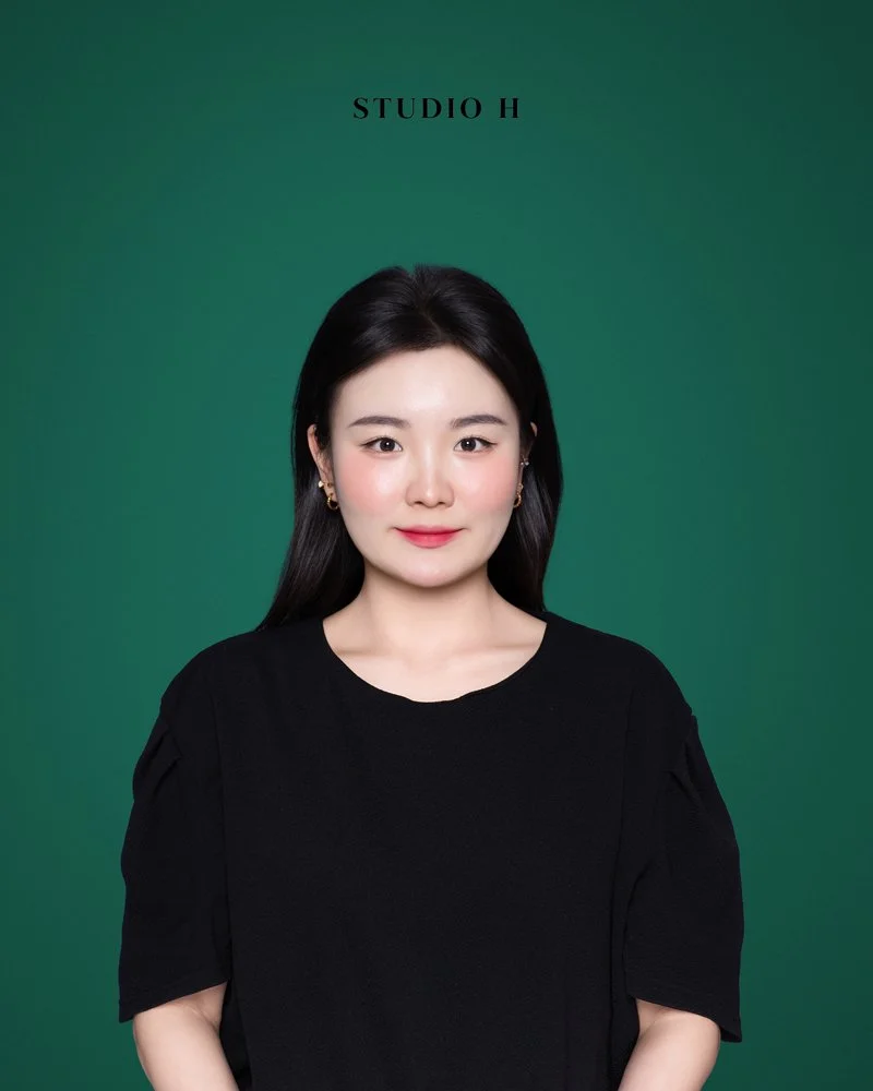 A woman with long black hair, wearing a black top and gold earrings, standing against a green background with 'STUDIO H' written above her head.