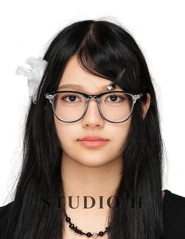 A young woman with long black hair wearing glasses, a black necklace, and hair clips, against a white background.