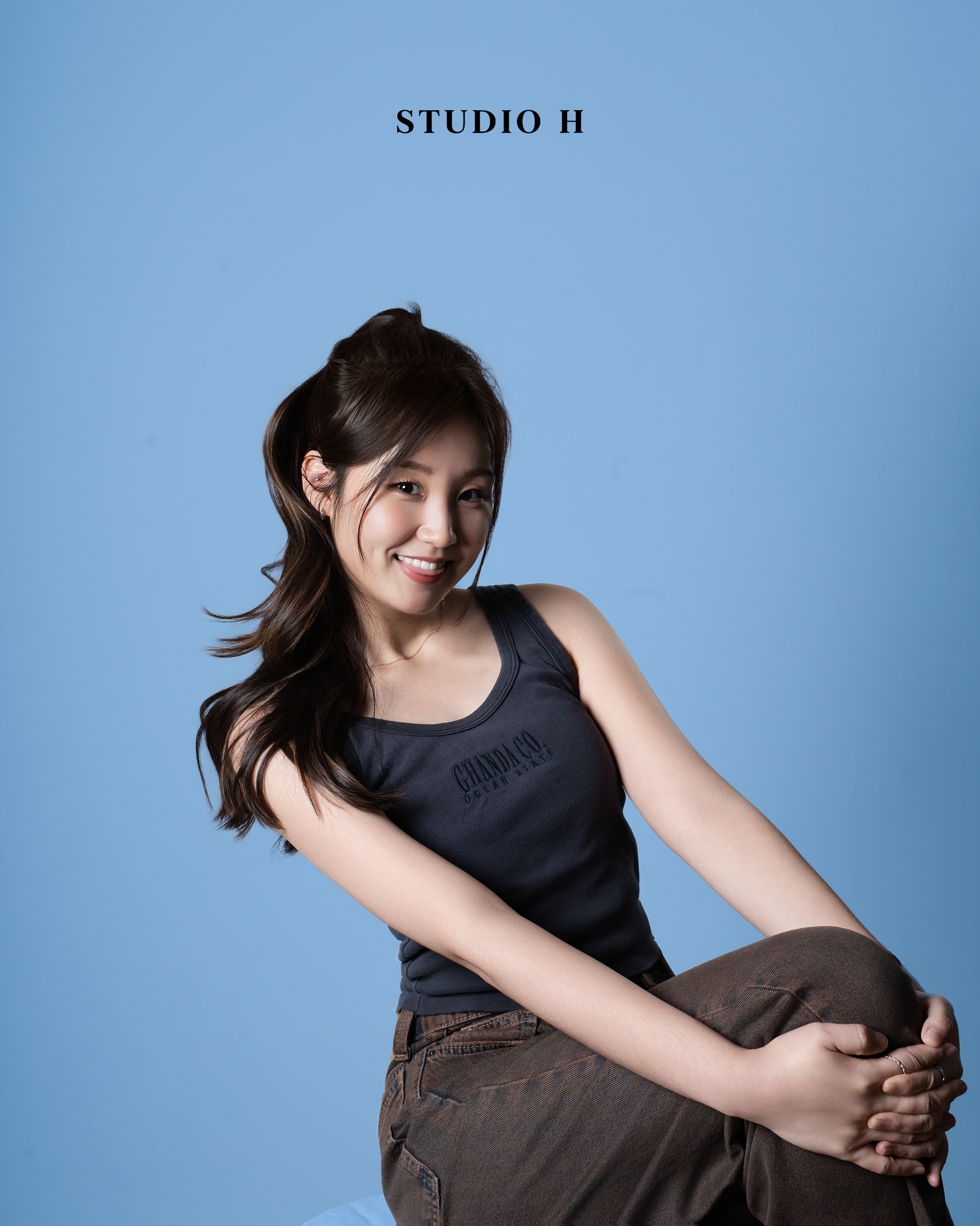 A young woman with long, wavy brown hair and a bright smile, wearing a dark tank top and brown pants, sitting against a light blue background with the text 'STUDIO H' at the top.