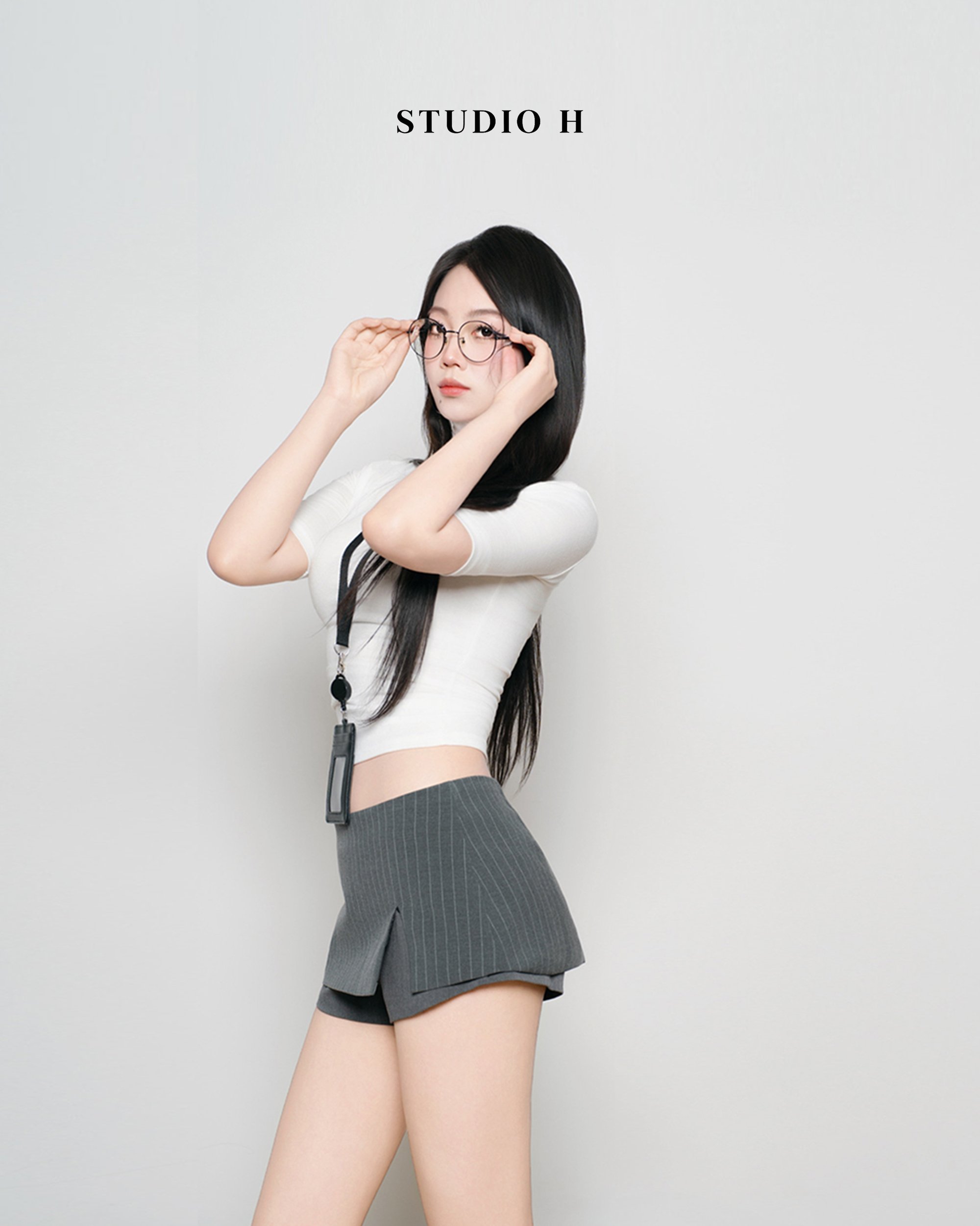 A young woman with long black hair and glasses adjusting her glasses, wearing a white crop top, gray pinstripe shorts, and a lanyard with an ID badge, standing against a plain white background with the text 'STUDIO H' above her.