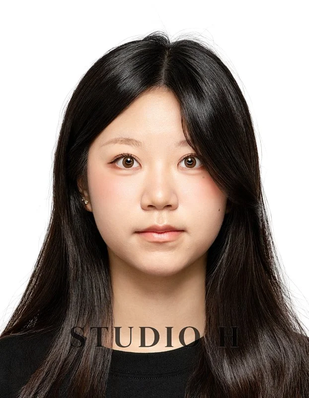 A portrait of a young Asian woman with long black hair, wearing a black top, looking directly at the camera against a plain white background.