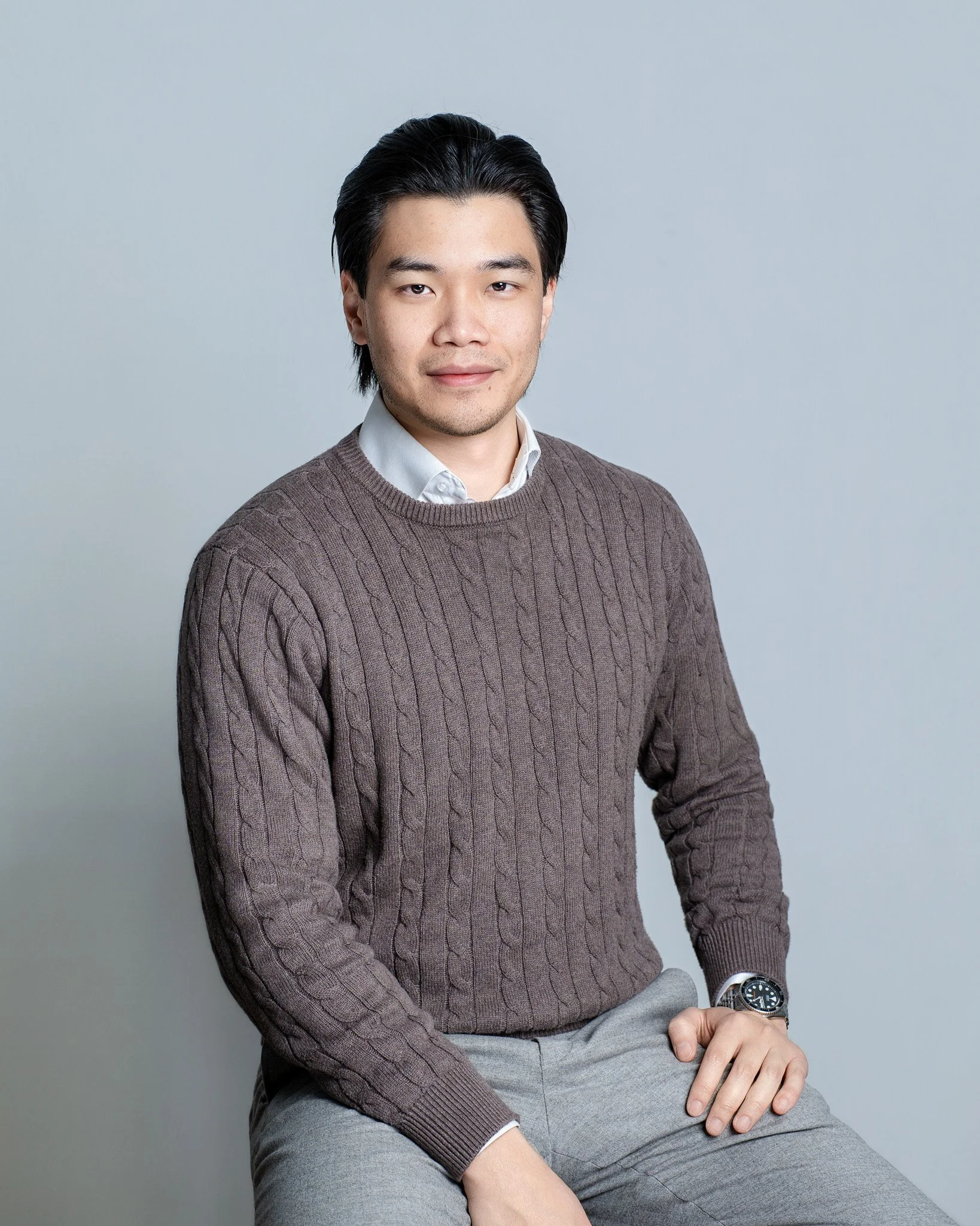 A young man with black hair wearing a white shirt, brown sweater, and gray pants, sitting against a plain light blue background, smiling slightly and looking at the camera.