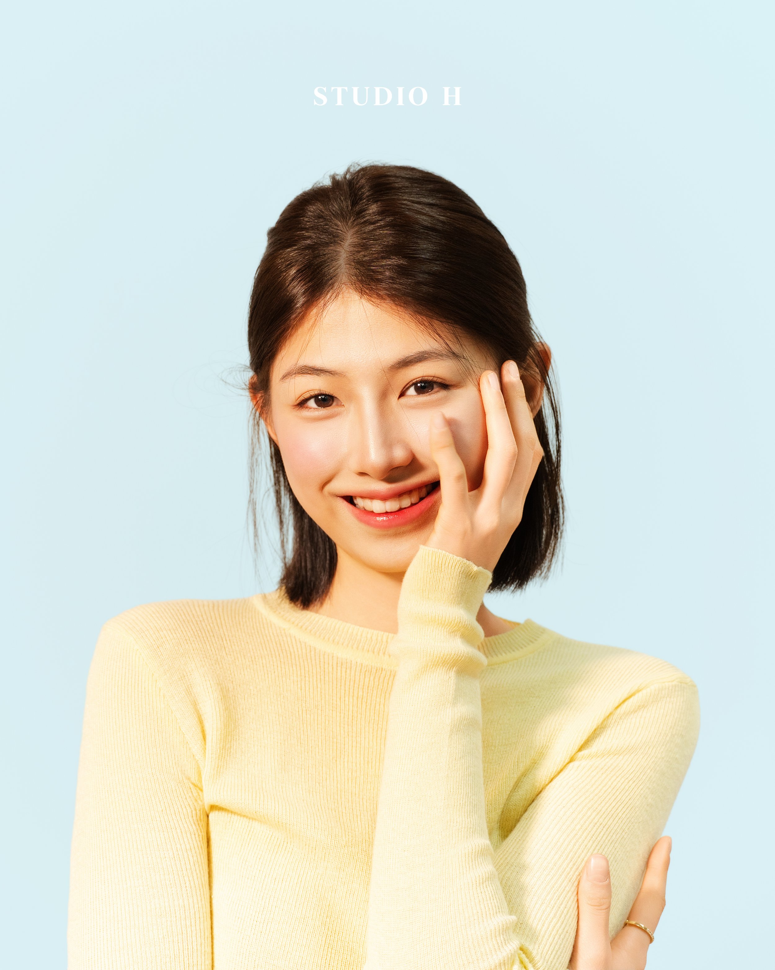 A young woman with dark hair smiling, winking, and touching her face with her right hand, wearing a yellow sweater, against a light blue background with 'STUDIO H' text at the top.