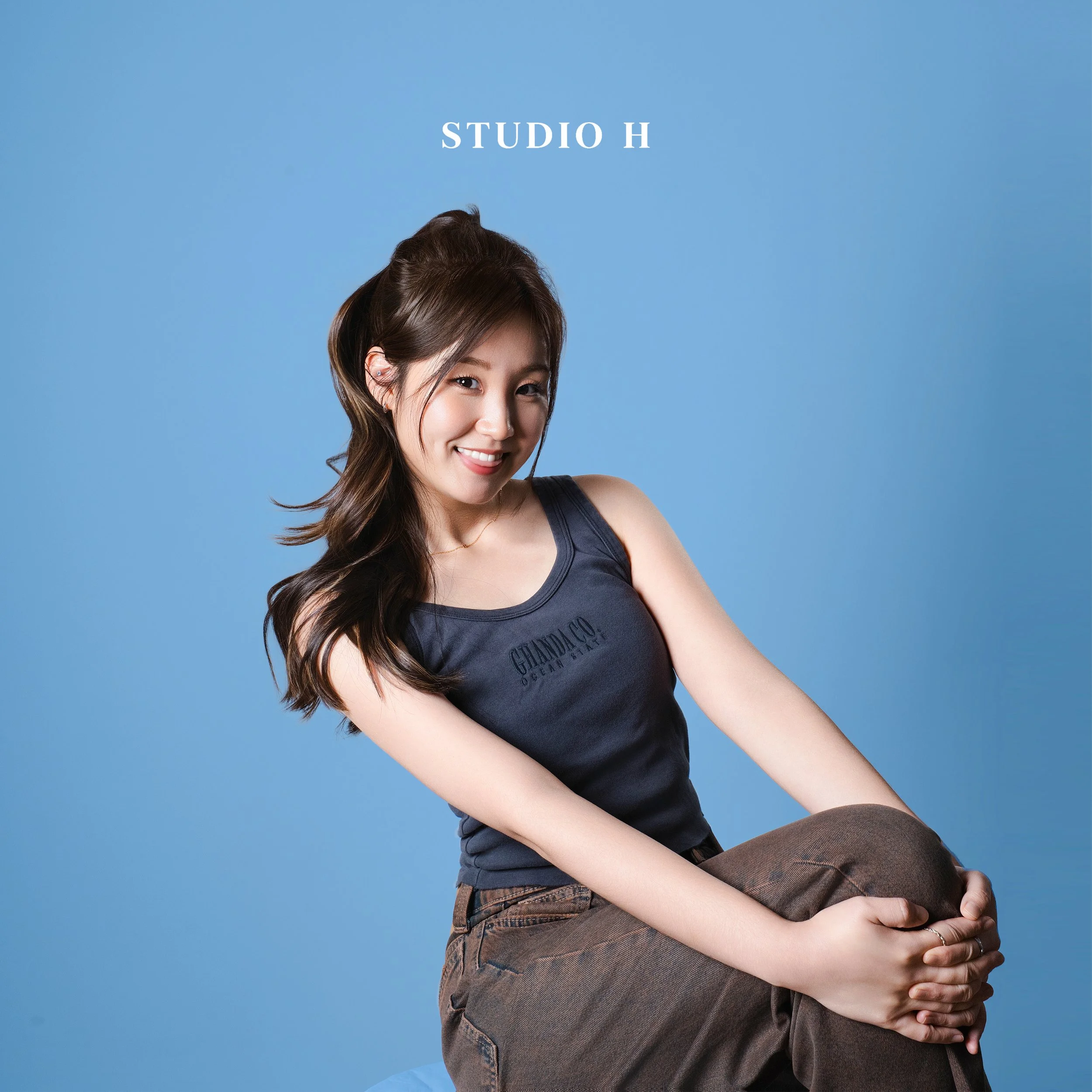 Close-up of a woman with long brown hair smiling against a blue background, wearing a gray tank top and brown pants.