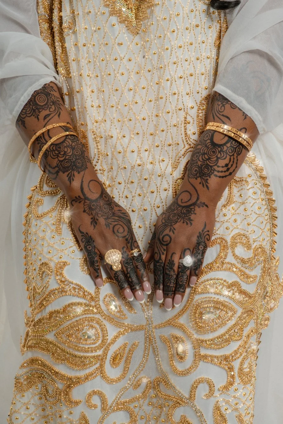 Person wearing a white garment with gold embroidery and pearls, adorned with gold rings and bracelets, with hands decorated in black henna intricate patterns.