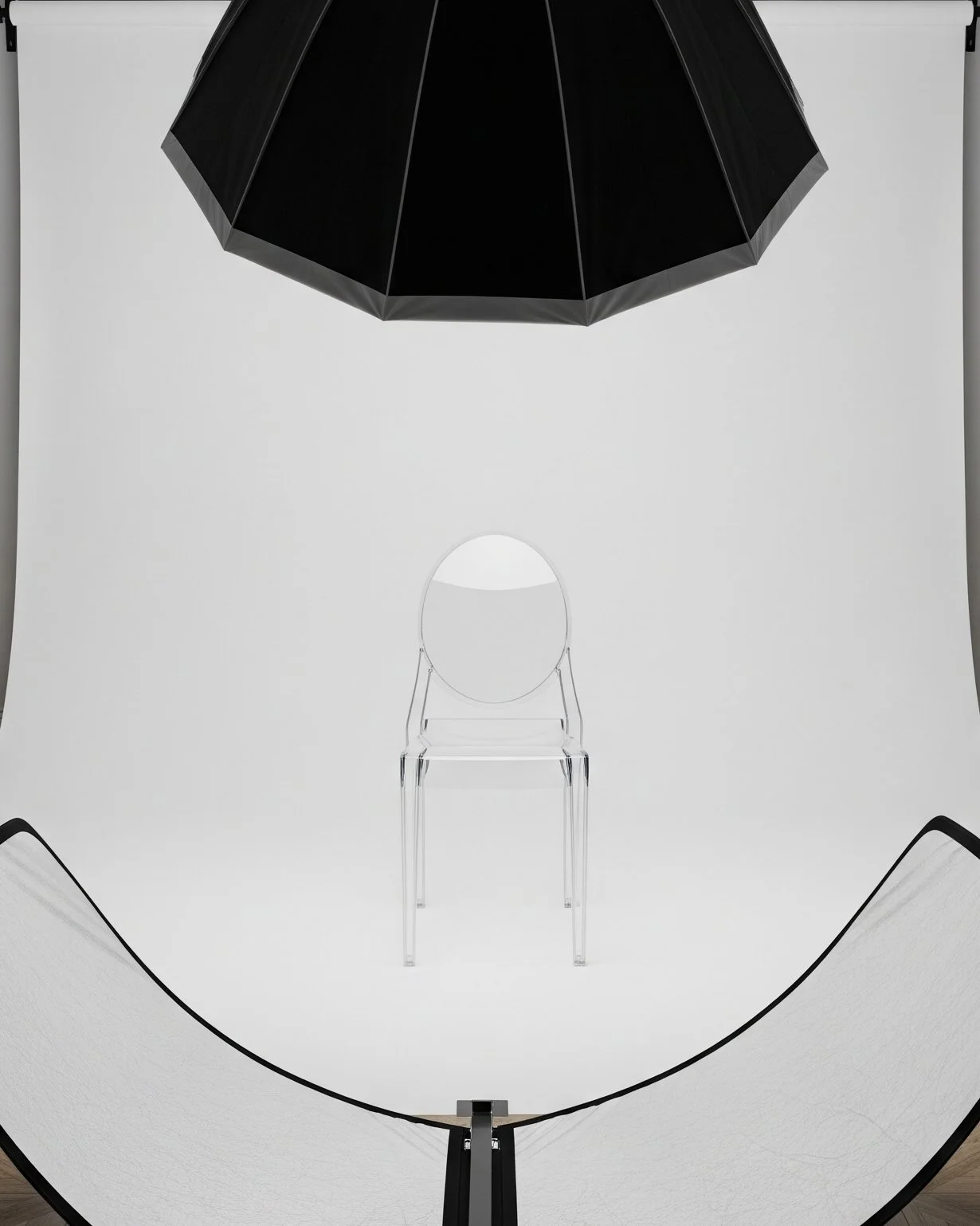 Photography studio setup with a clear plastic chair centered in front of a white backdrop, a large black umbrella-style light diffuser overhead, and two white reflectors or background supports on the sides.