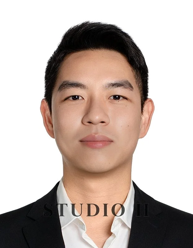 A professional headshot of a young man in a black suit and white shirt, smiling subtly, with a white background.
