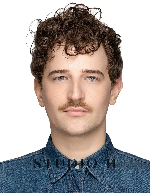 Portrait of a young man with curly brown hair, blue eyes, a mustache, wearing a denim shirt.