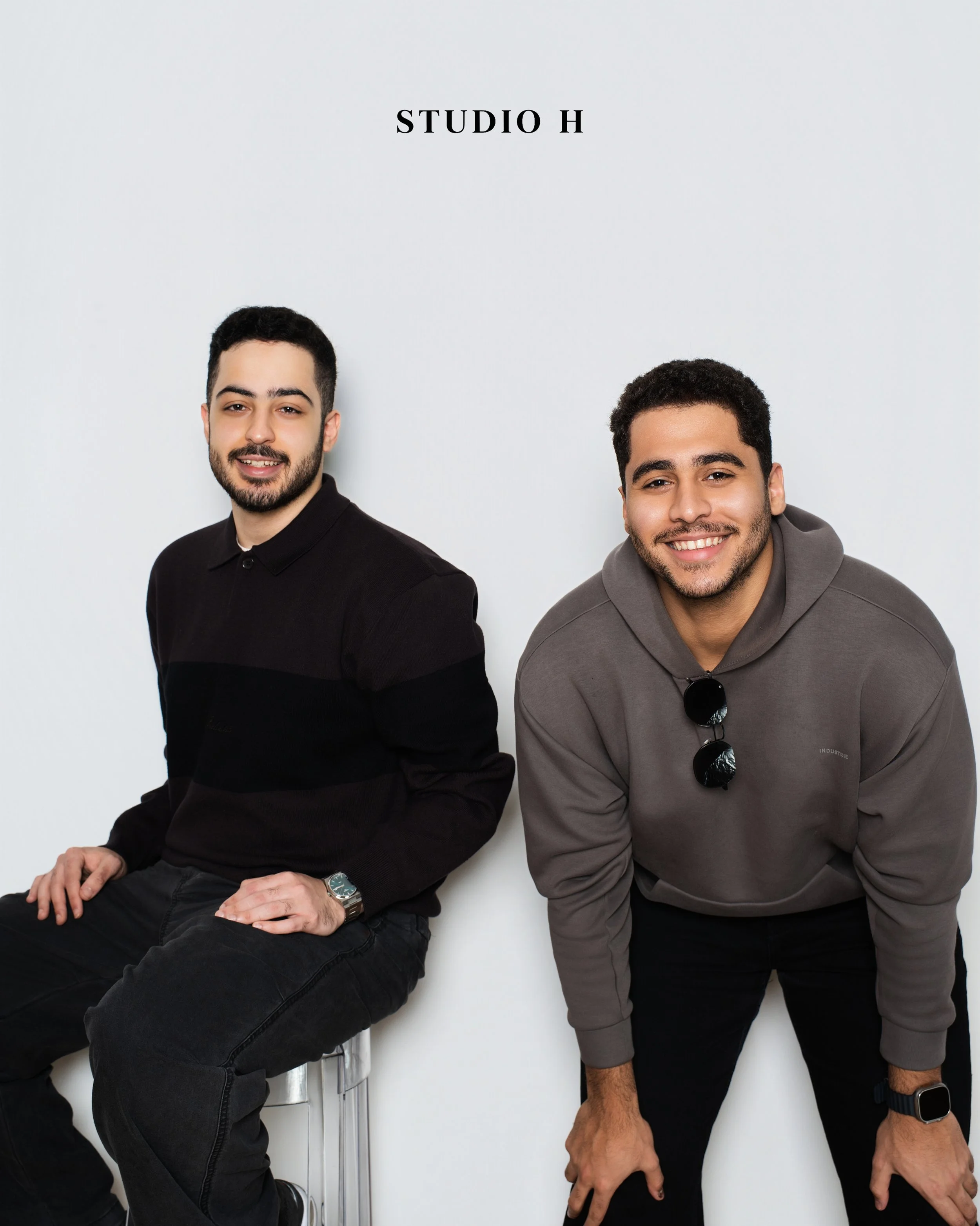 Two young men posing against a white wall with 'STUDIO H' written above them. One is sitting and the other is leaning forward, both smiling.