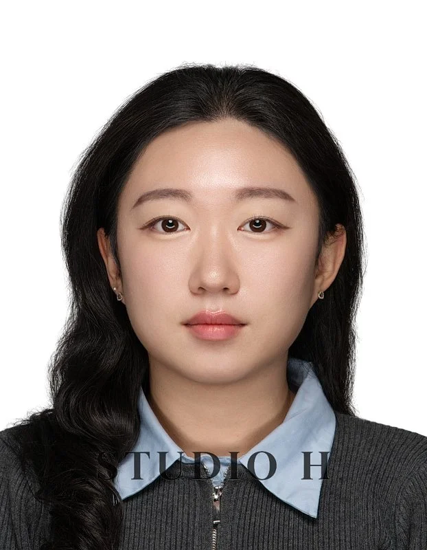 A young woman with long black hair styled in loose waves, wearing a blue collared shirt under a black zip-up sweater, looking directly at the camera with a neutral expression, against a white background.