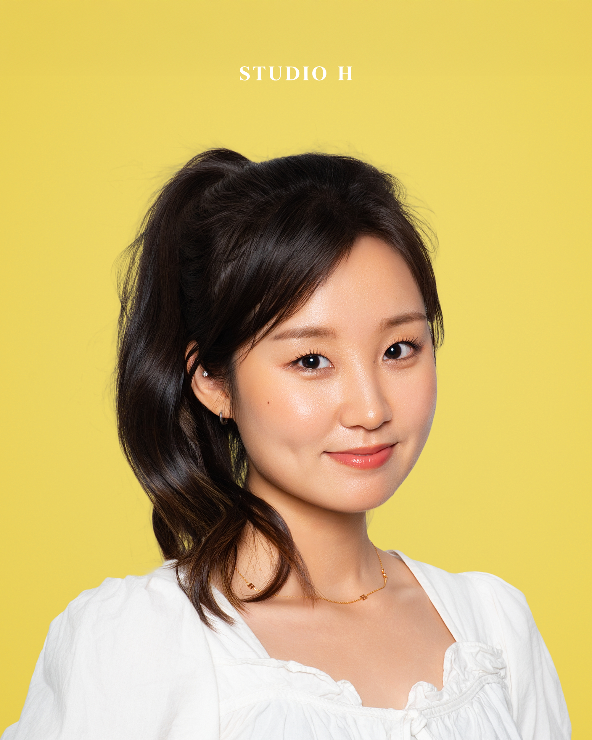 A young woman with dark hair styled to the side, wearing a white top and a delicate gold necklace, smiling softly against a yellow background with the text 'STUDIO H' at the top.